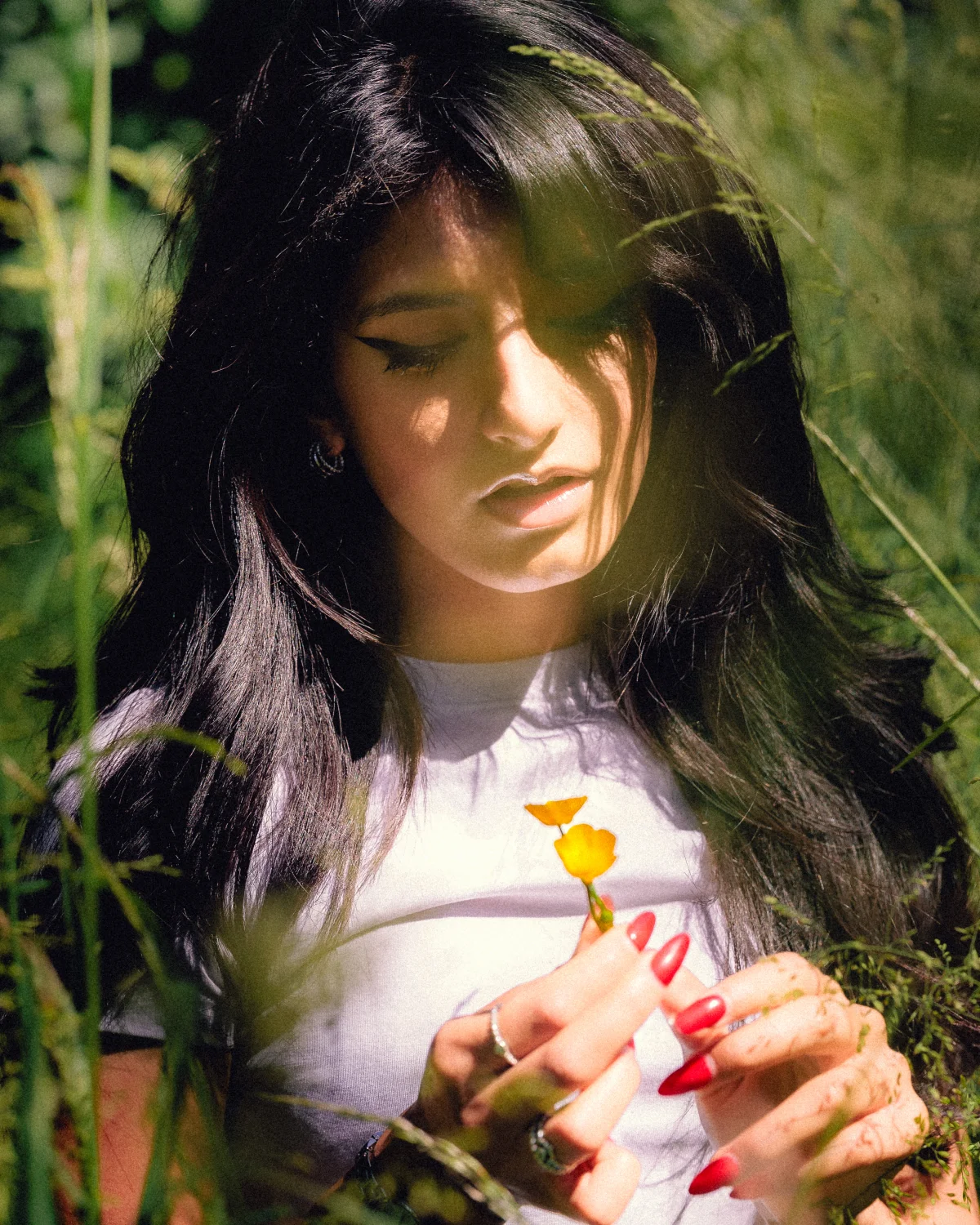 Young woman with long dark hair looking down at a yellow flower in a sunny field.