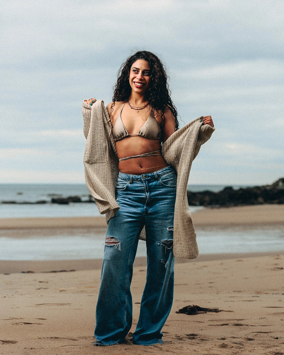 A smiling woman with curly hair posing on a sandy beach in a bikini top, wide-leg jeans, and an open cardigan.