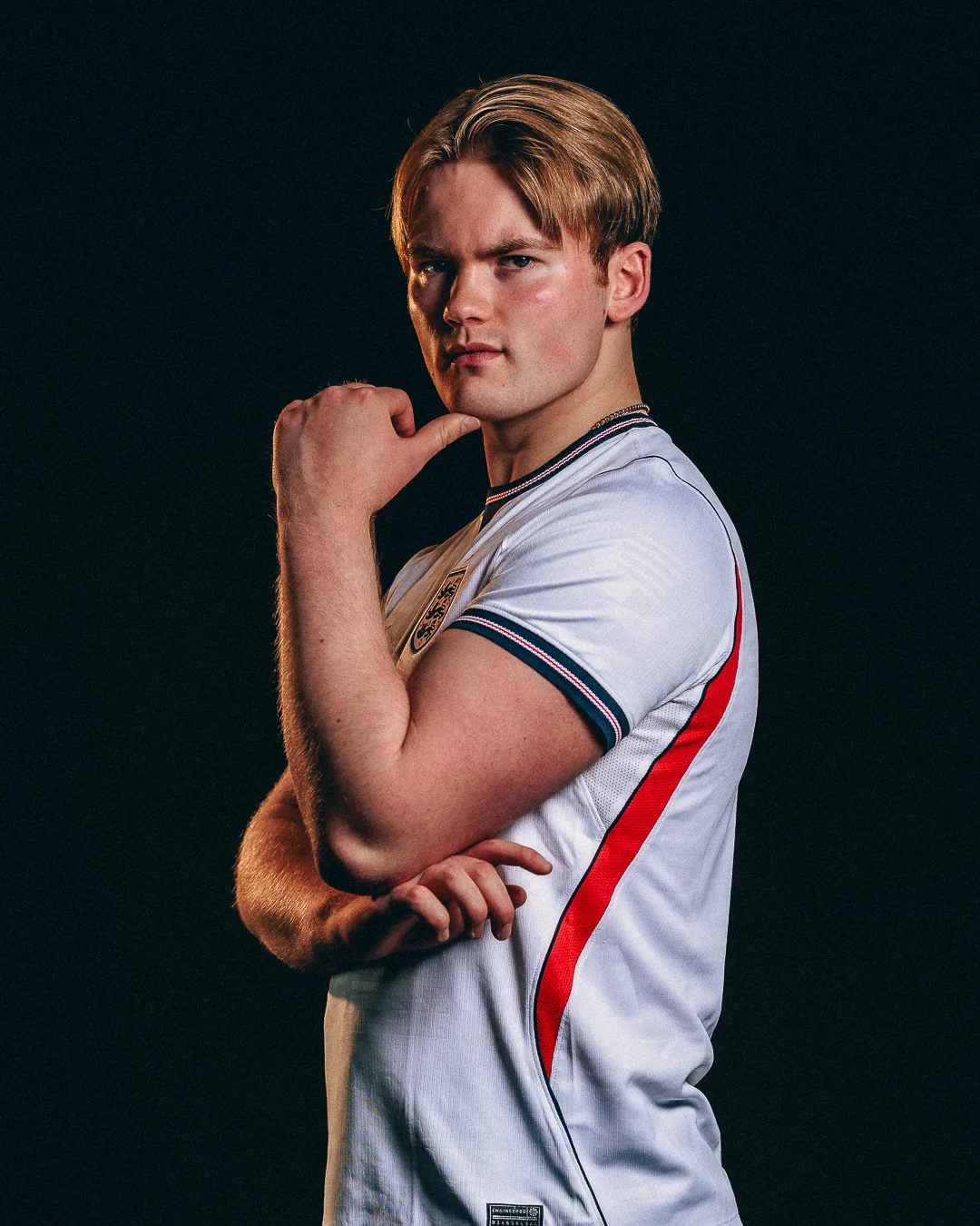 A young man with blond hair in a white sports jersey posing thoughtfully against a solid black background.