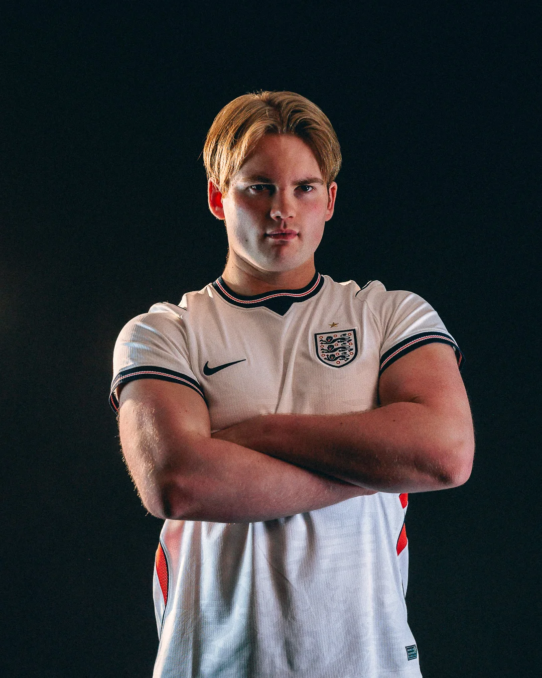 A serious young man in an England football jersey with arms crossed against a dark studio background.