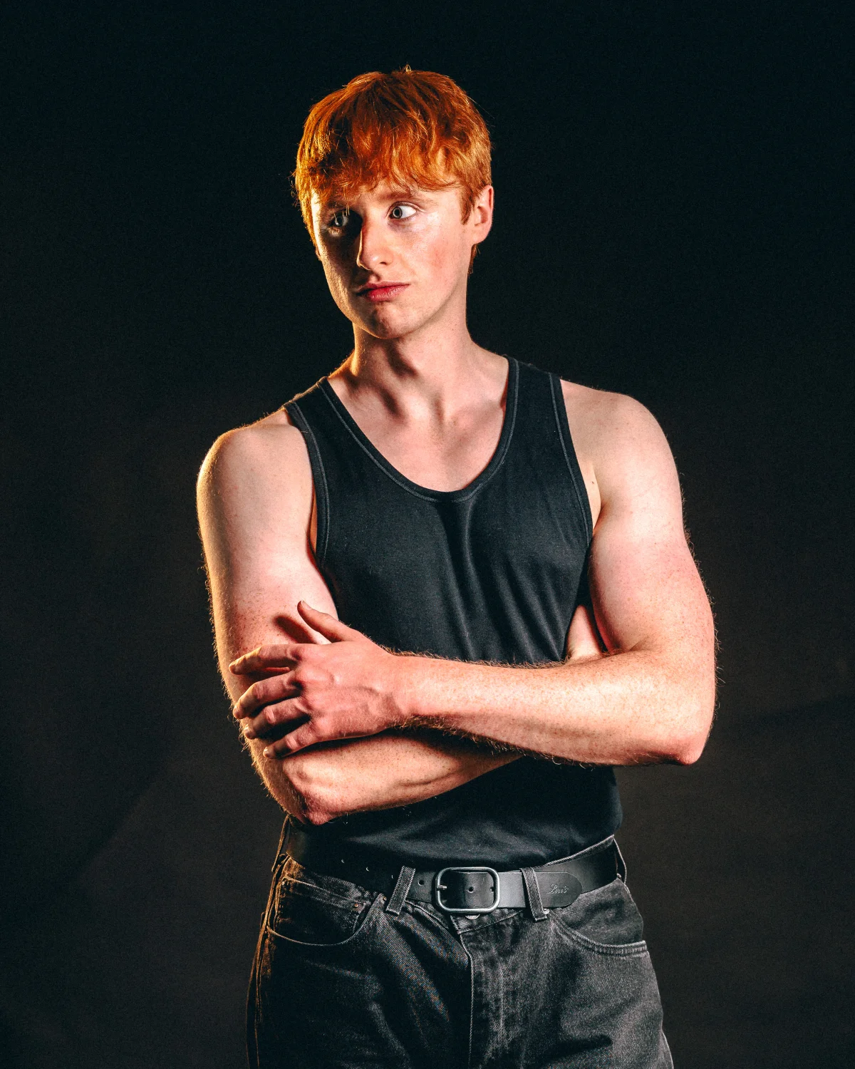 Studio portrait of a red-haired man in a black tank top with arms crossed against a dark background.