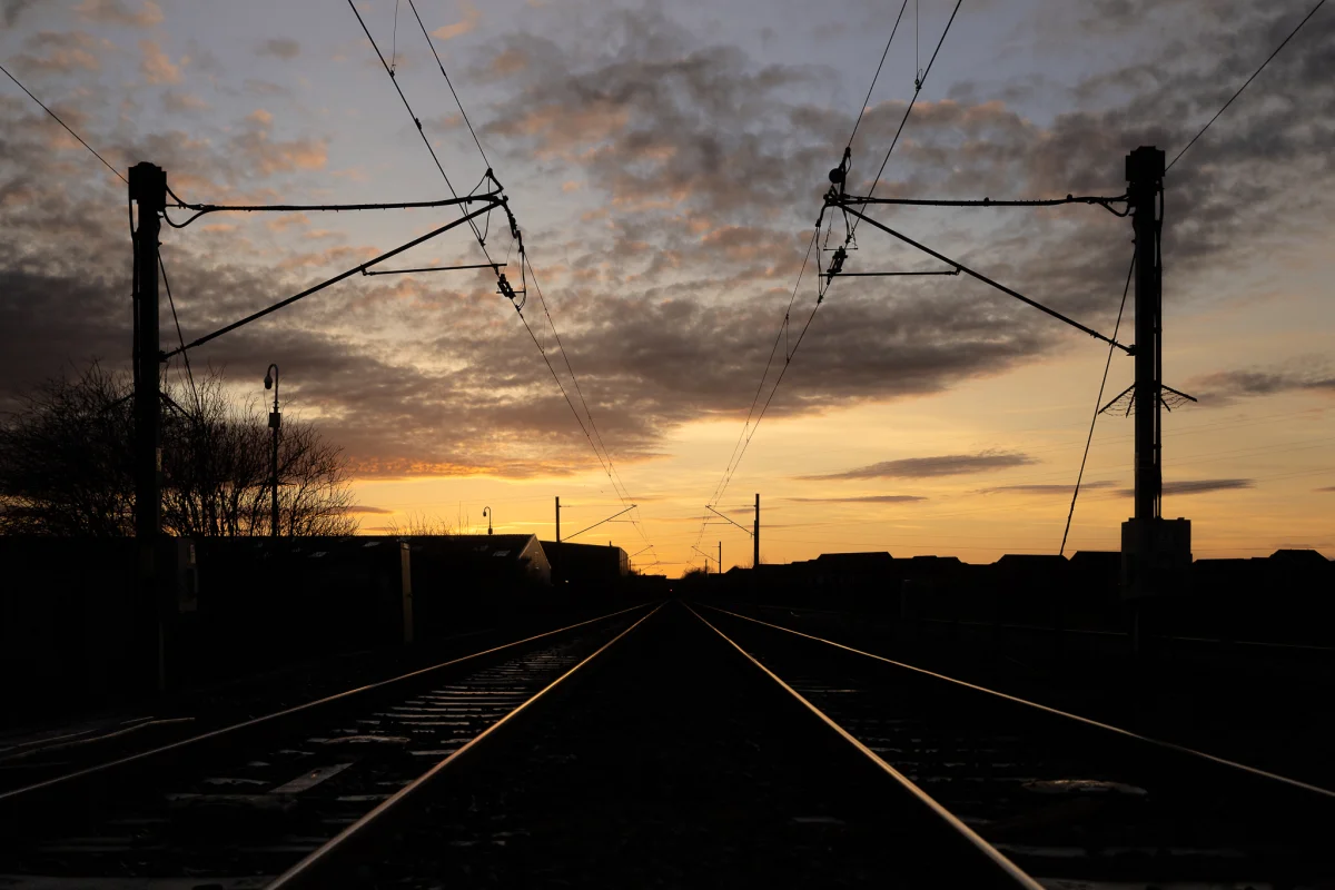 Silhouette of railway tracks and overhead power lines leading toward a glowing sunset sky.