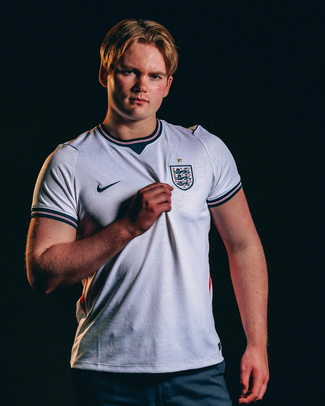 A young man with blonde hair wearing an England football jersey posing against a dark studio background.