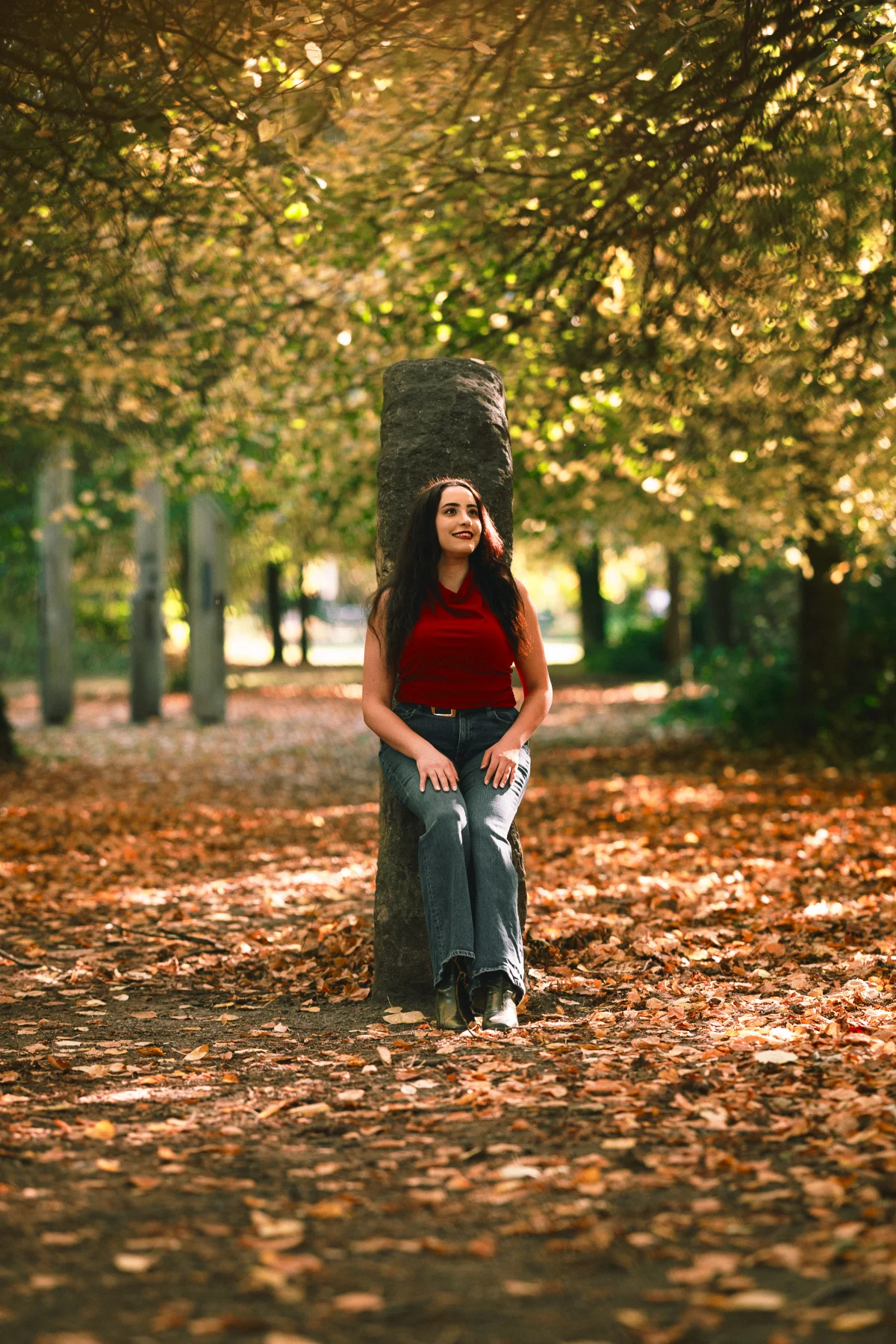 A woman in a red top and blue jeans leans against a stone pillar in an autumn park with fallen leaves.