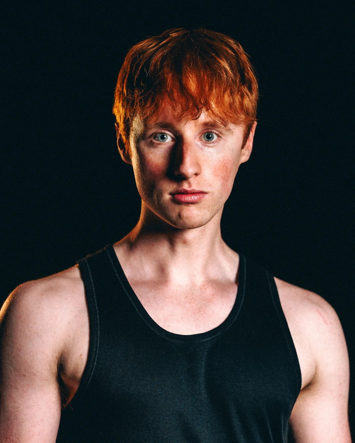Close-up portrait of a young man with red hair and blue eyes wearing a black tank top against a dark background.