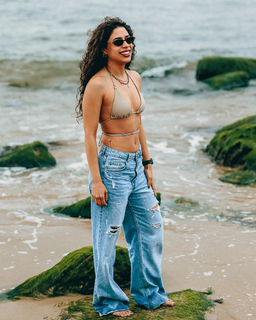 A smiling woman posing on a rocky beach wearing a beige bikini top and ripped blue jeans.