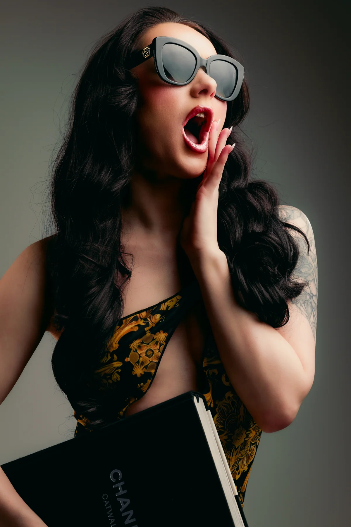 A woman with long dark hair and oversized black sunglasses posing with a surprised expression in a floral dress.