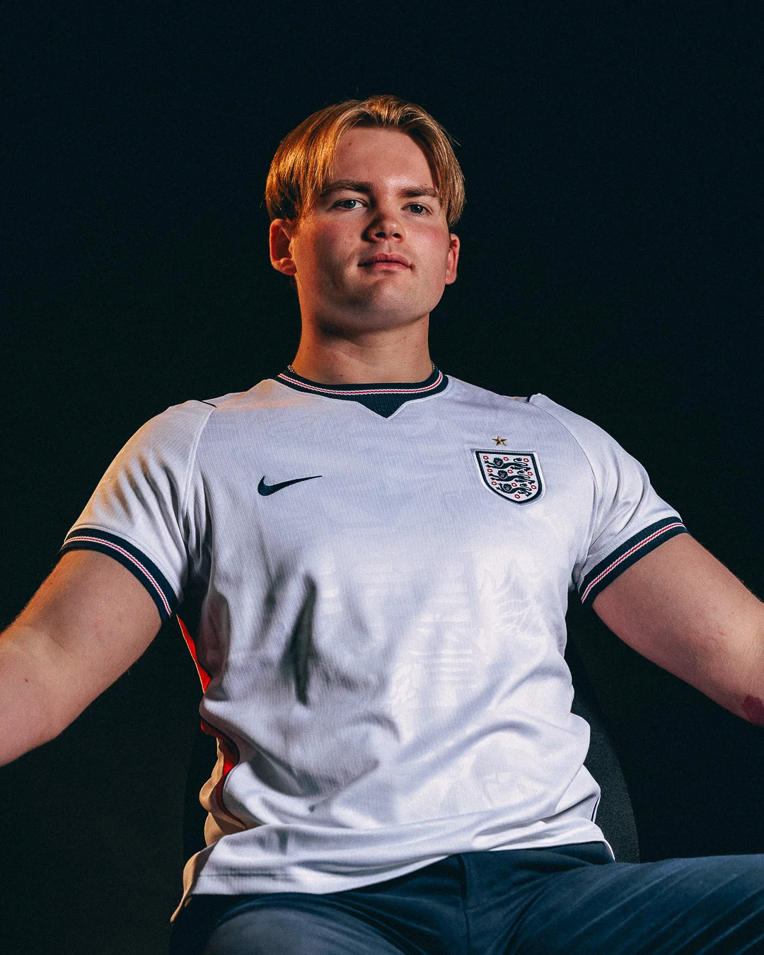 Young man in a white England football jersey against a dark background with dramatic studio lighting.