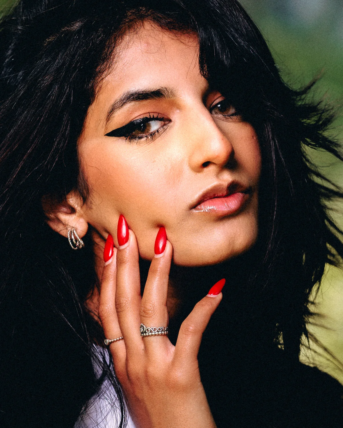 Close-up portrait of a woman with bold winged eyeliner and red nails against a blurred green background.