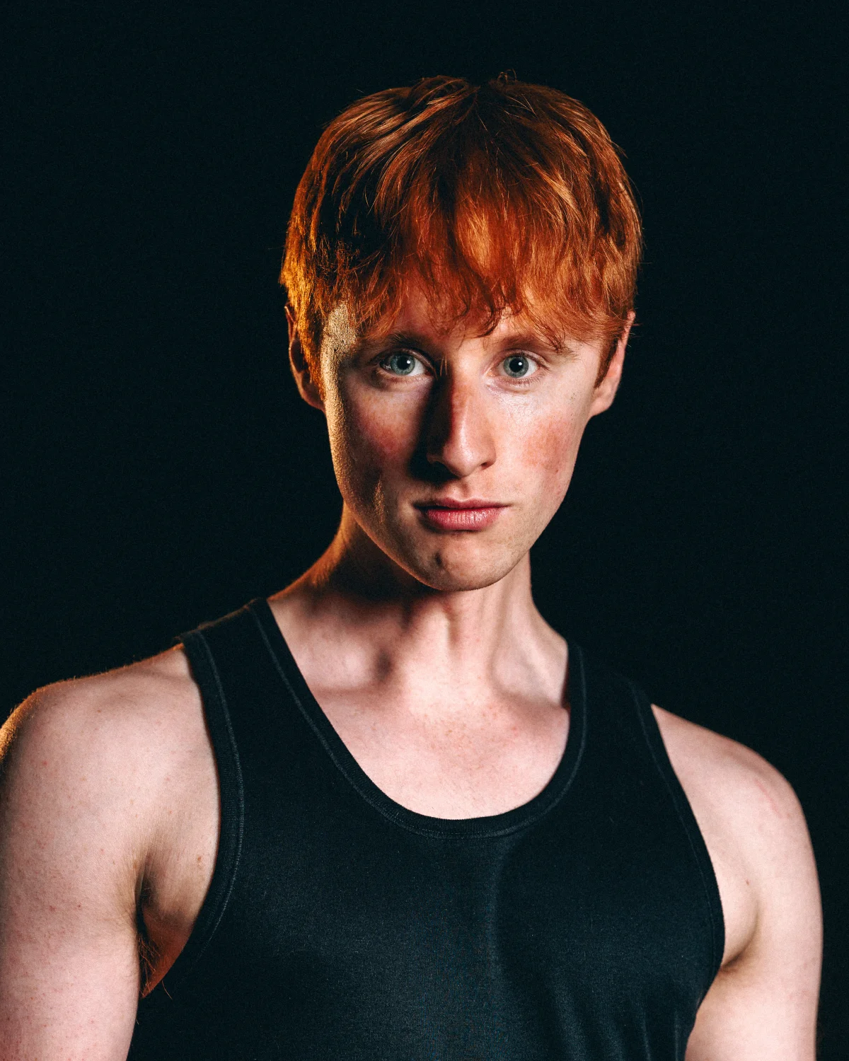Studio portrait of a man with red hair wearing a black tank top against a solid black background.