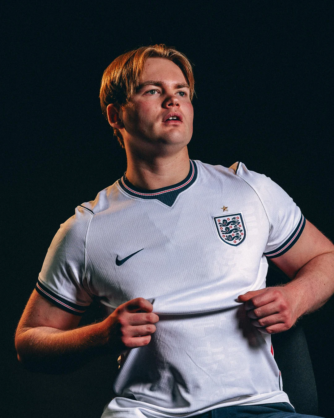 A portrait of a young man in an England football jersey against a dark studio background.
