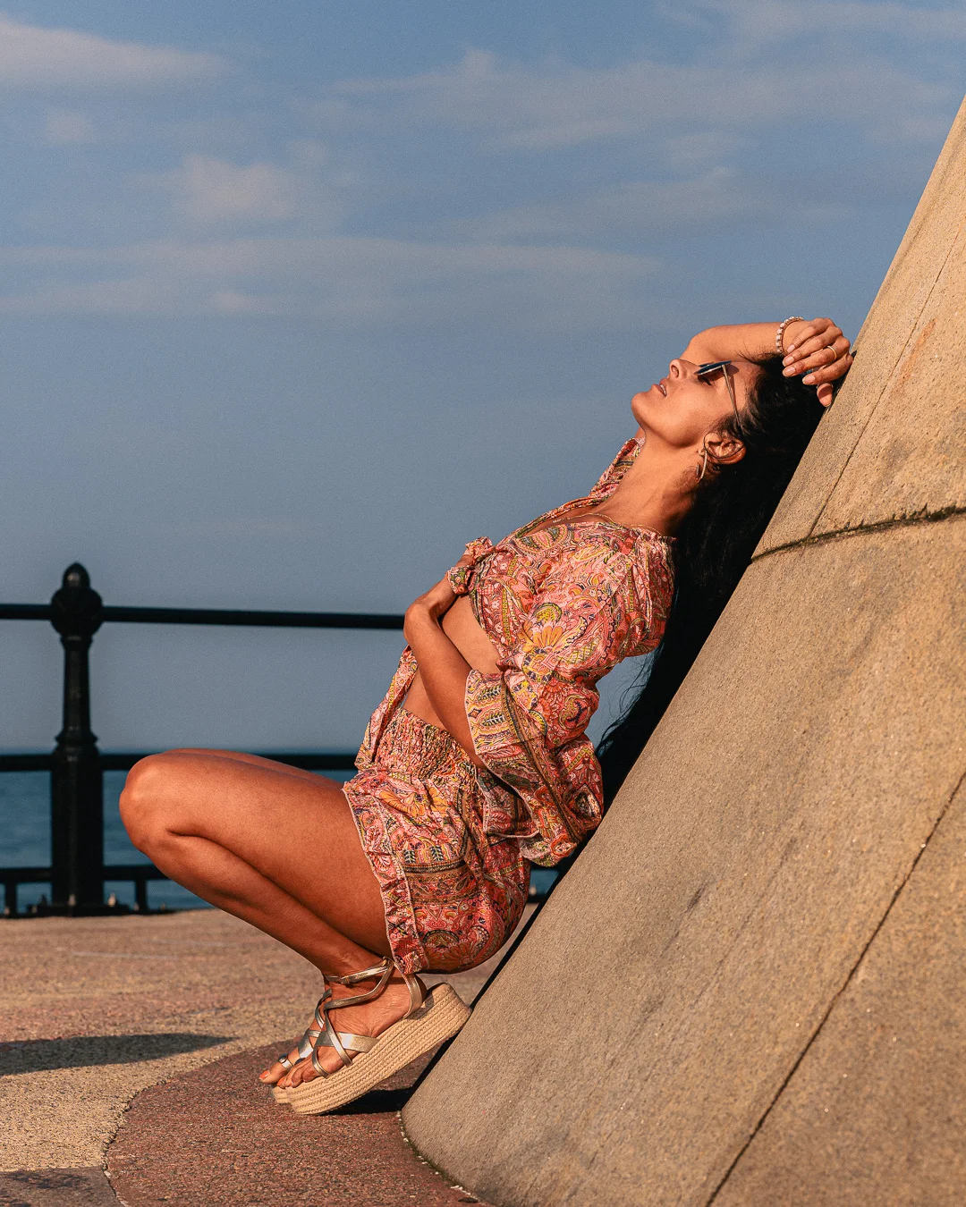 Woman in a colorful patterned two-piece outfit leaning against a concrete wall by the ocean at sunset.