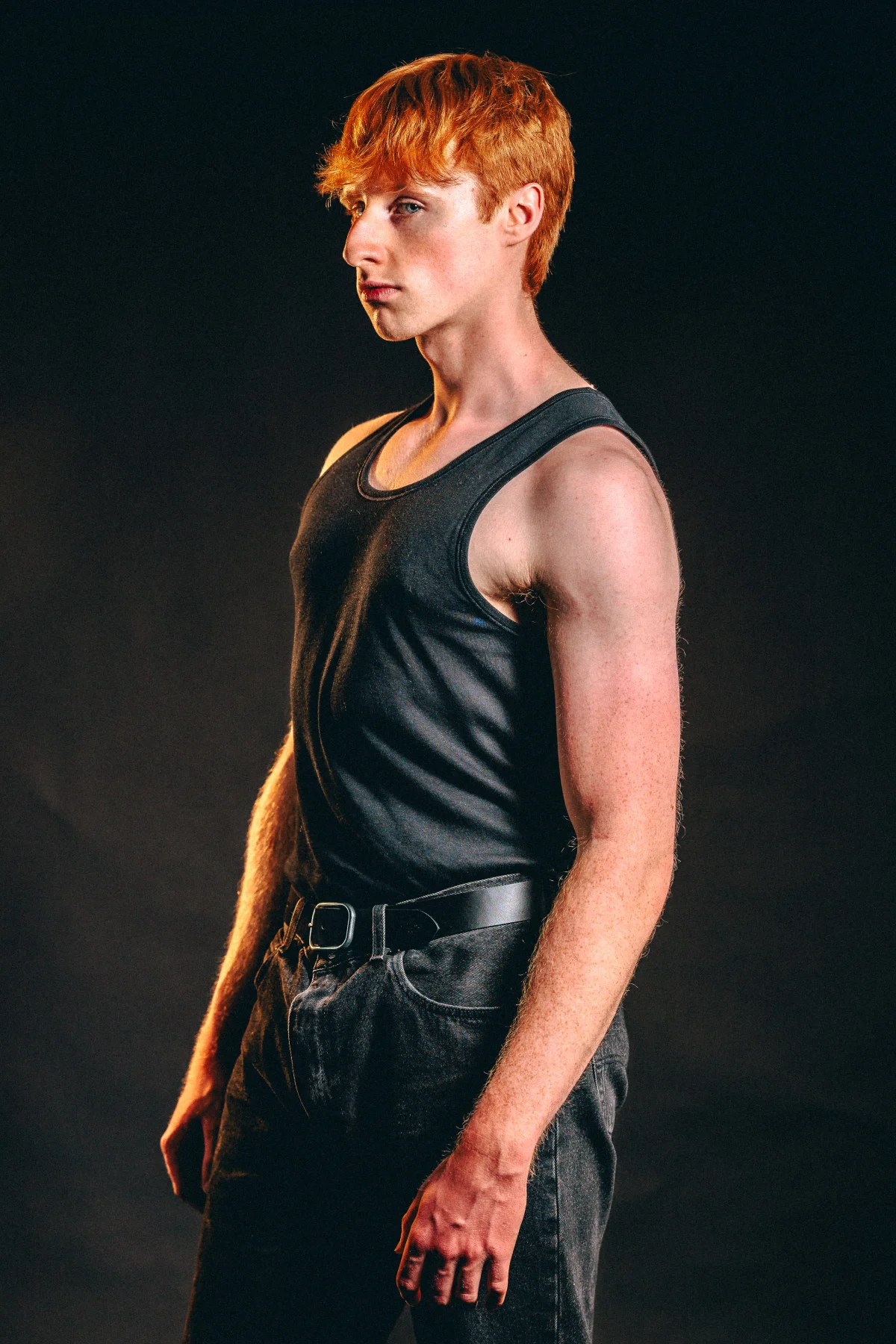 A young man with red hair in a black tank top and dark jeans posing against a black background.