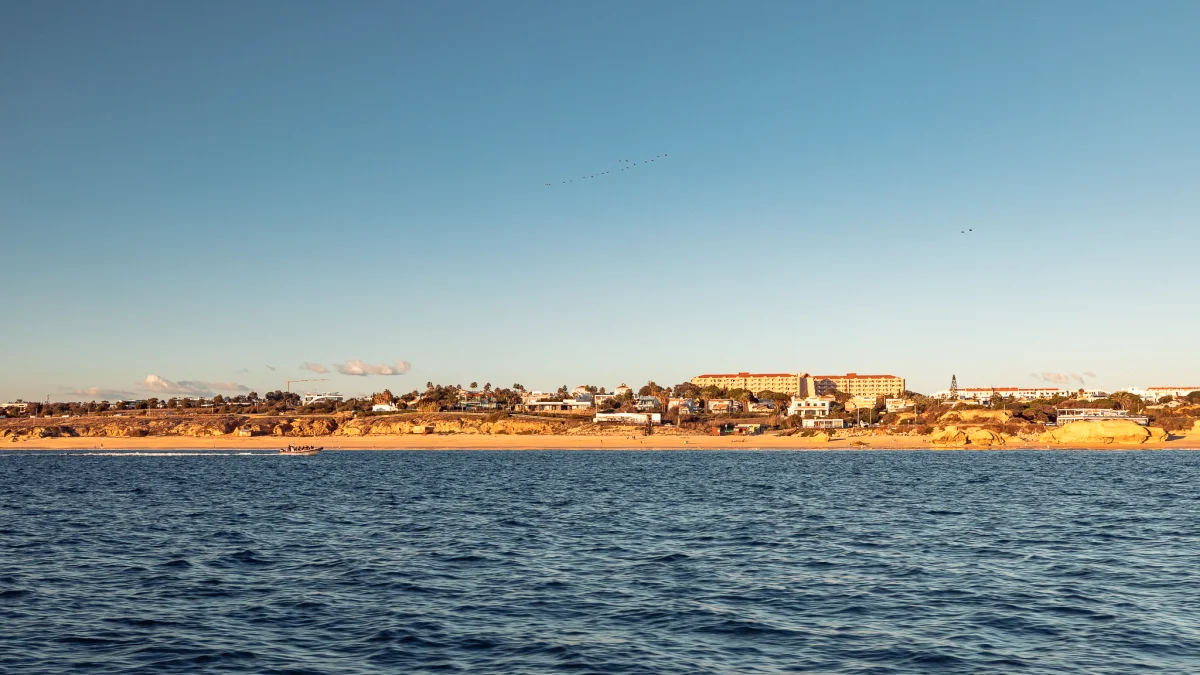 Wide shot of a coastal town with sandy cliffs and buildings overlooking a calm blue ocean under a clear sky.