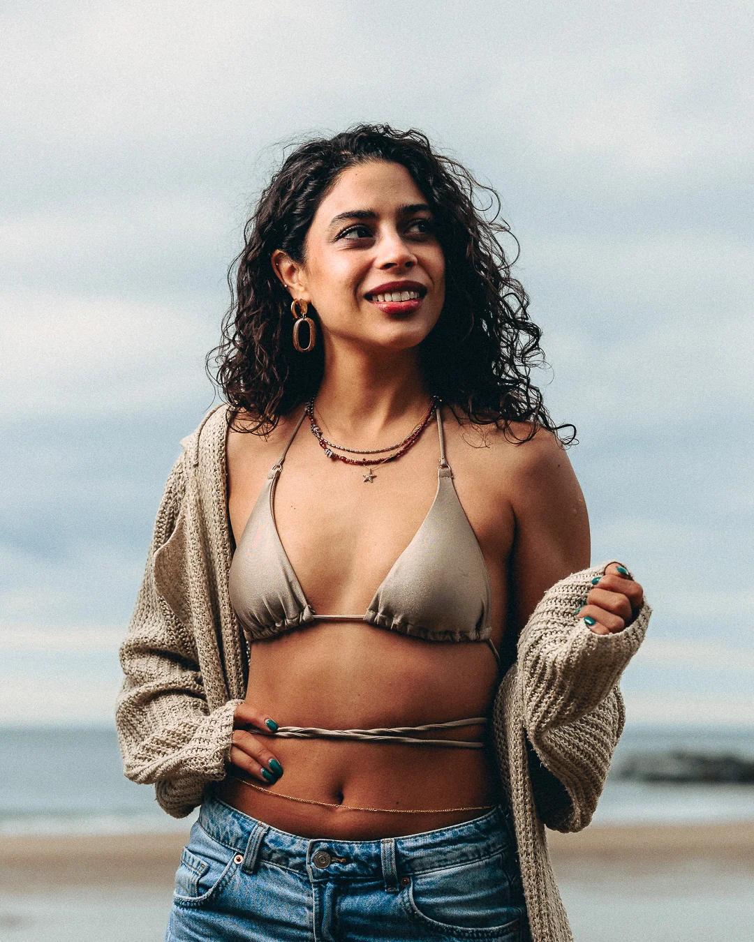 A woman with curly hair posing on a beach wearing a beige bikini top, denim shorts, and an open cardigan.