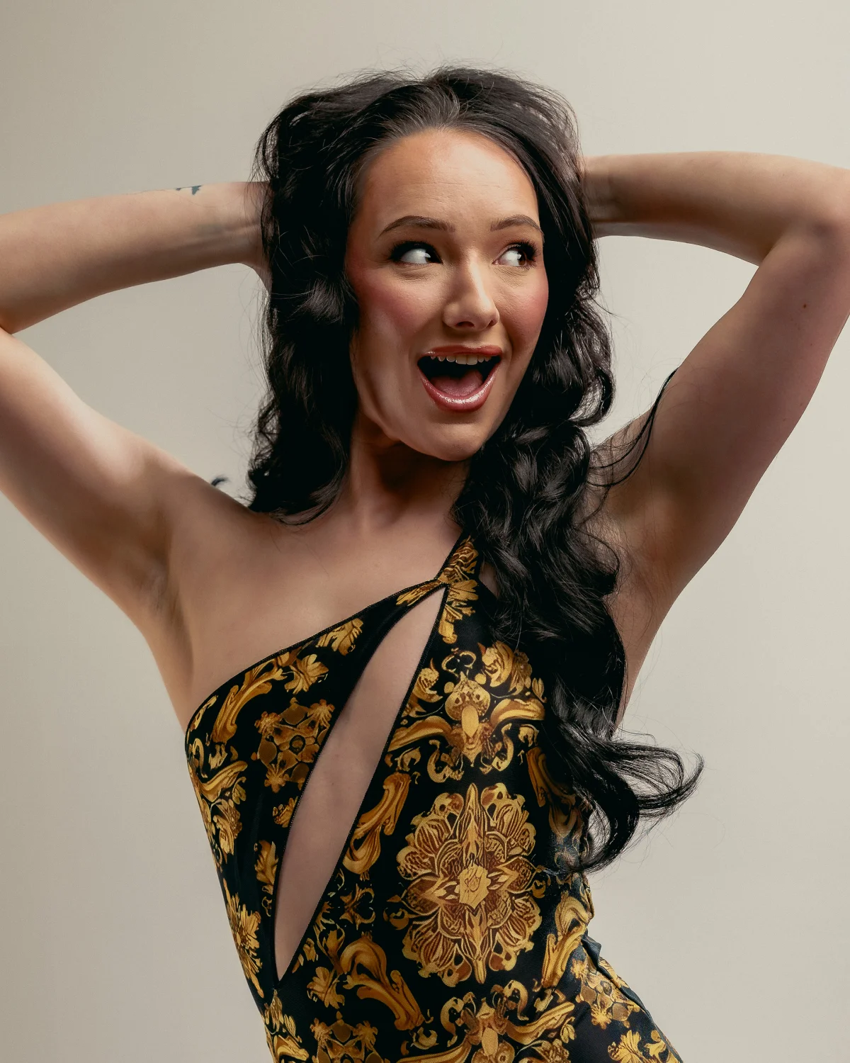 A woman with long dark wavy hair and an excited expression wearing a gold and black patterned one-shoulder dress.