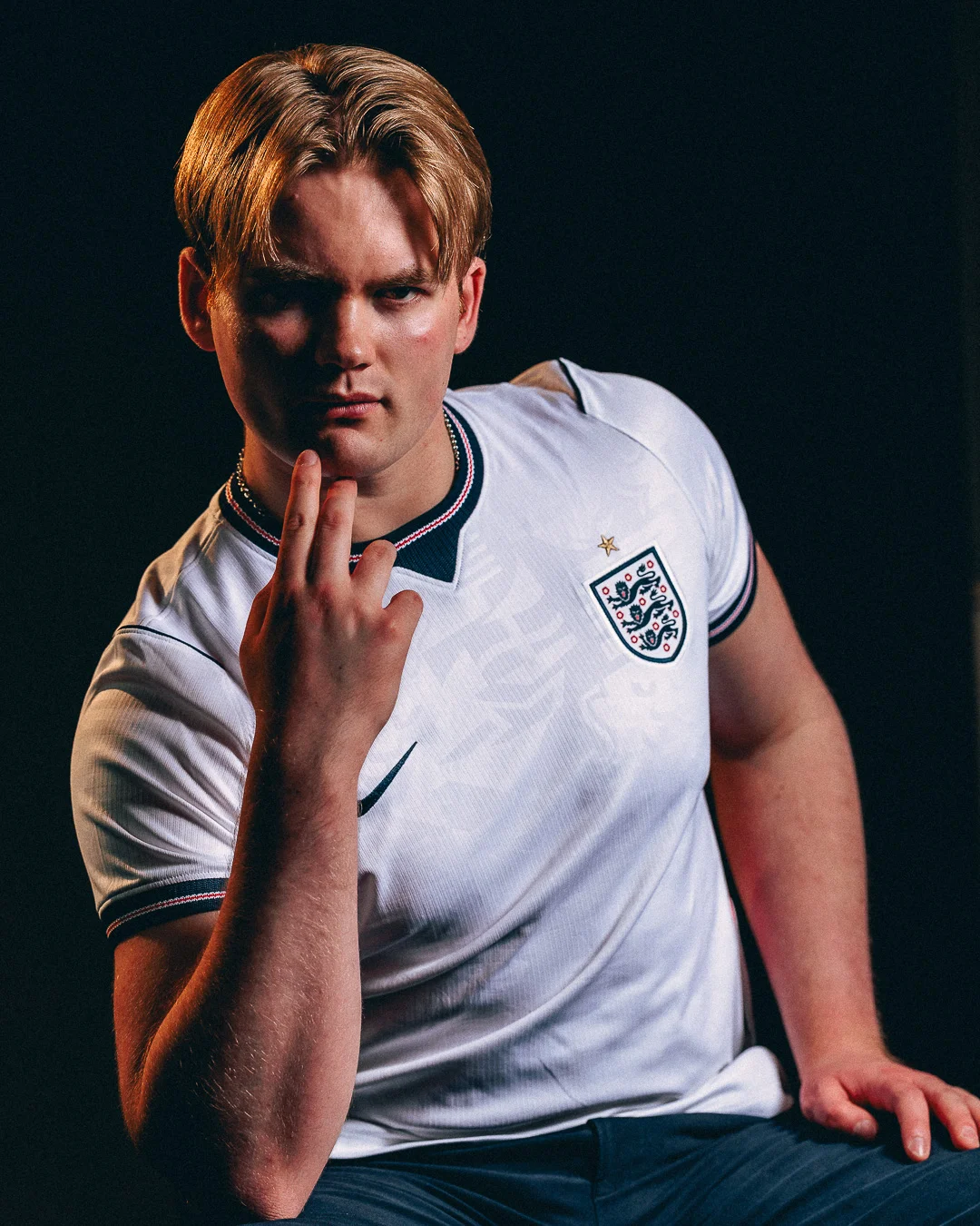 A moody studio portrait of a young man with blonde hair wearing an England football jersey.