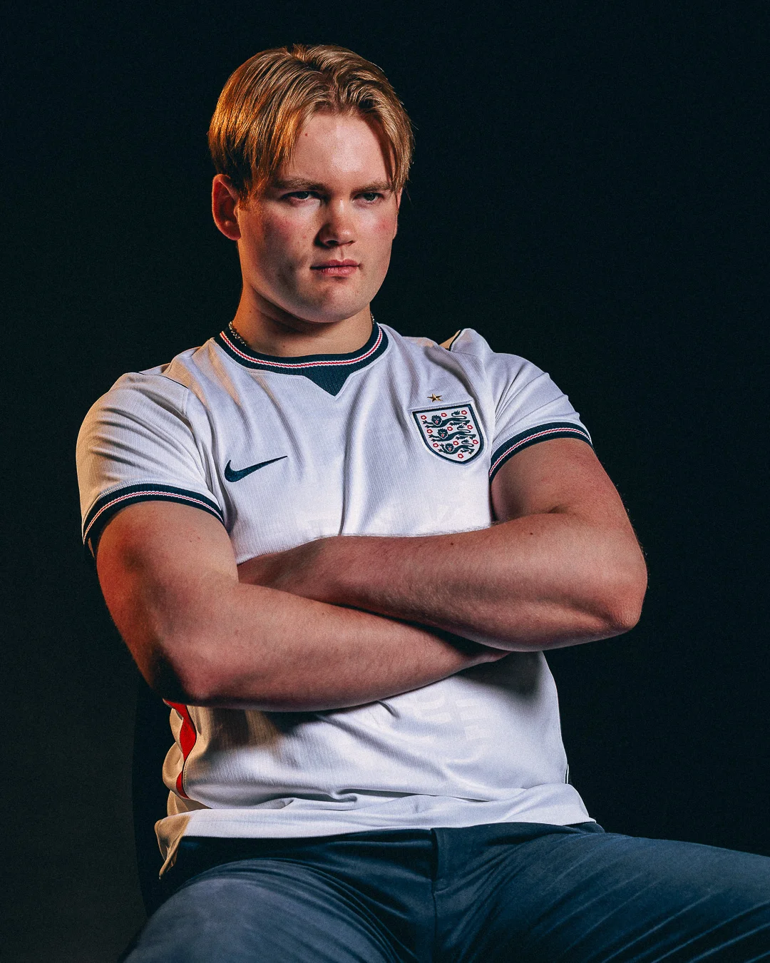 Young man with blonde hair in an England football jersey, arms crossed against a dark background.