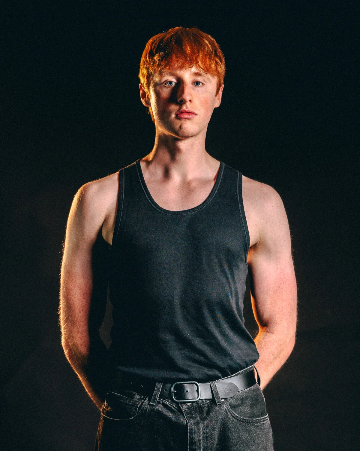 A young man with red hair wearing a black tank top and jeans, posing against a dark background with moody lighting.