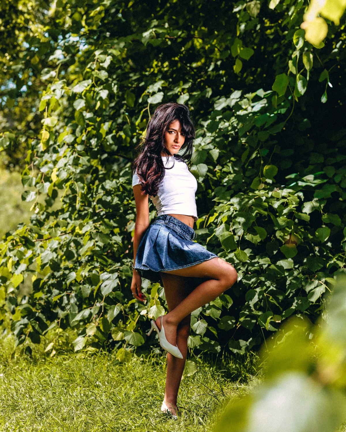 Woman with long dark hair posing in a white t-shirt and denim mini skirt against lush green foliage.