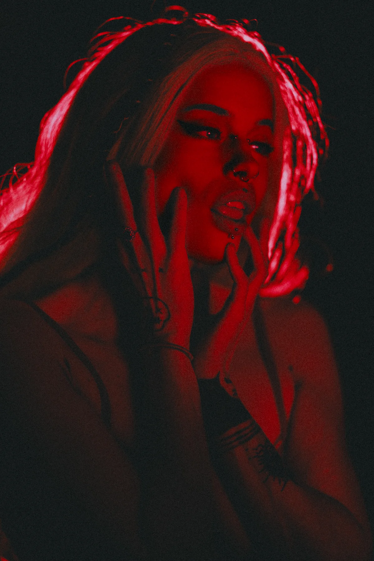 Woman with blonde hair in intense red lighting, hands framing her face against a black background.