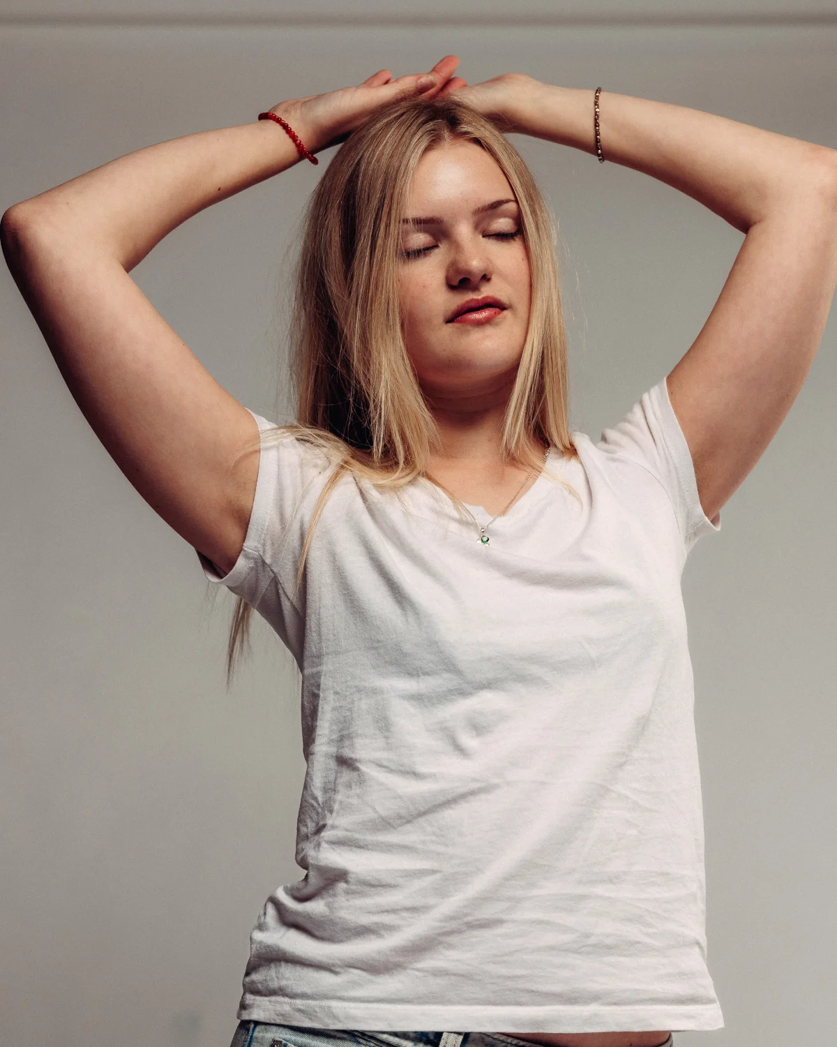 Portrait of a blonde woman in a white t-shirt with eyes closed and hands behind her head against a grey background.