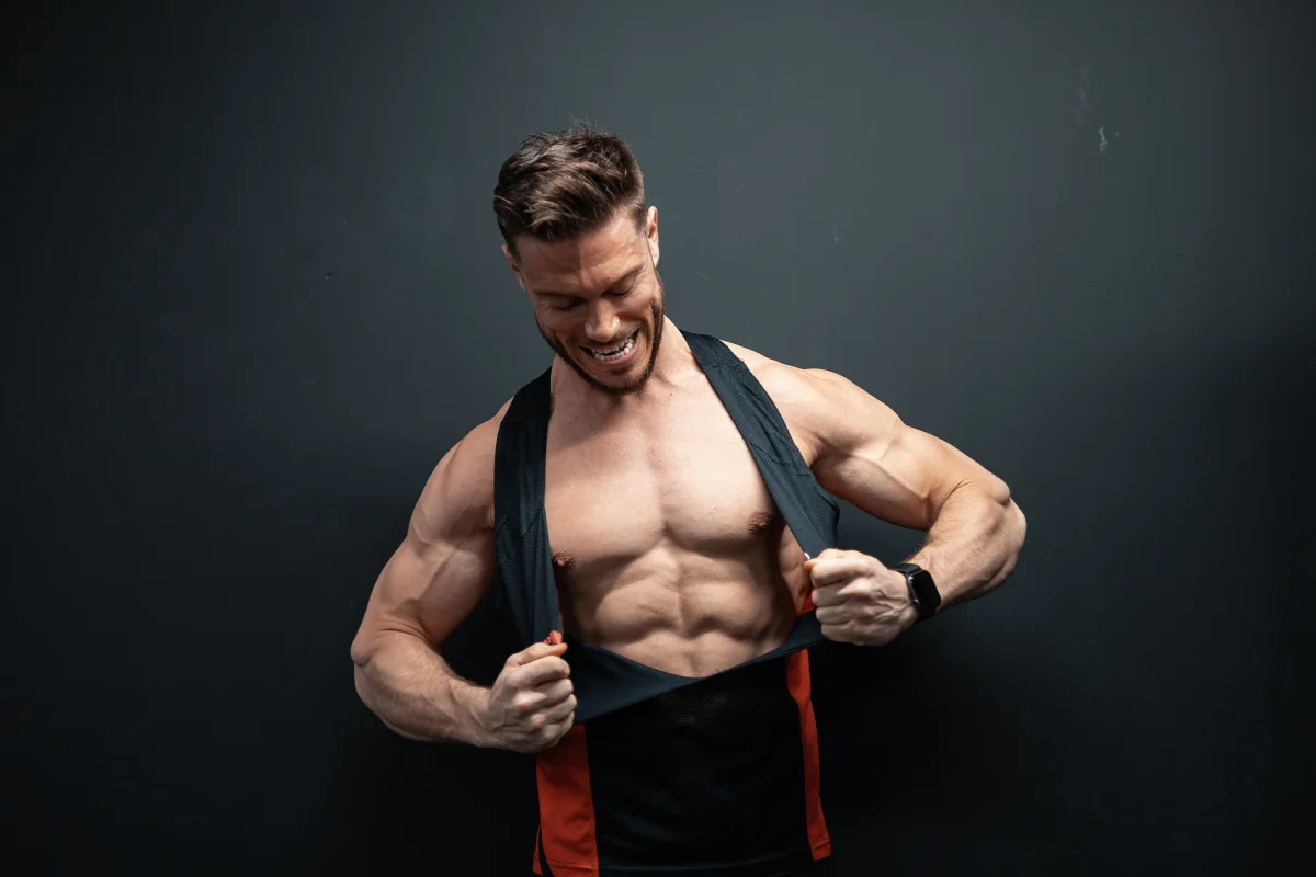 Muscular man smiling and pulling up his black tank top to show his chest and abs against a dark studio background.