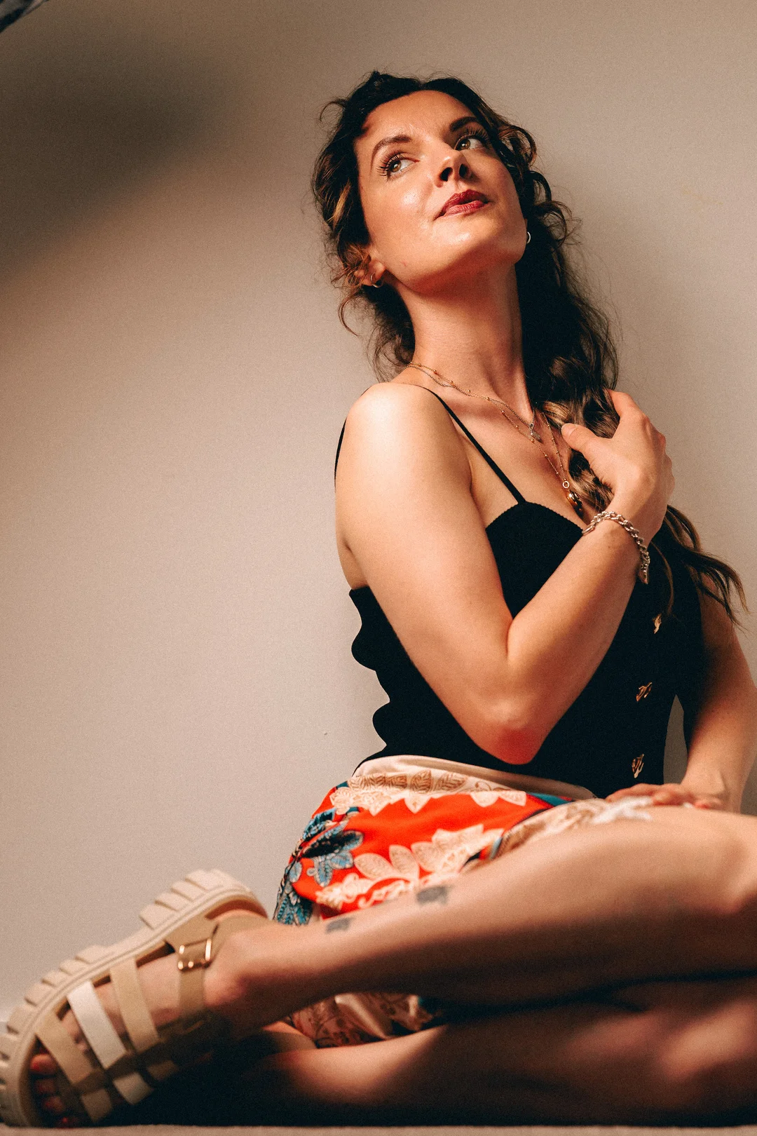 A woman with long brown hair in a black top and red patterned skirt, gazing upwards in a moody studio setting.