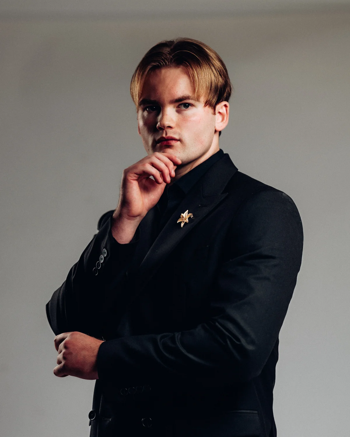 A young man with reddish-blonde hair in a black suit and gold lapel pin, posing thoughtfully against a grey background.