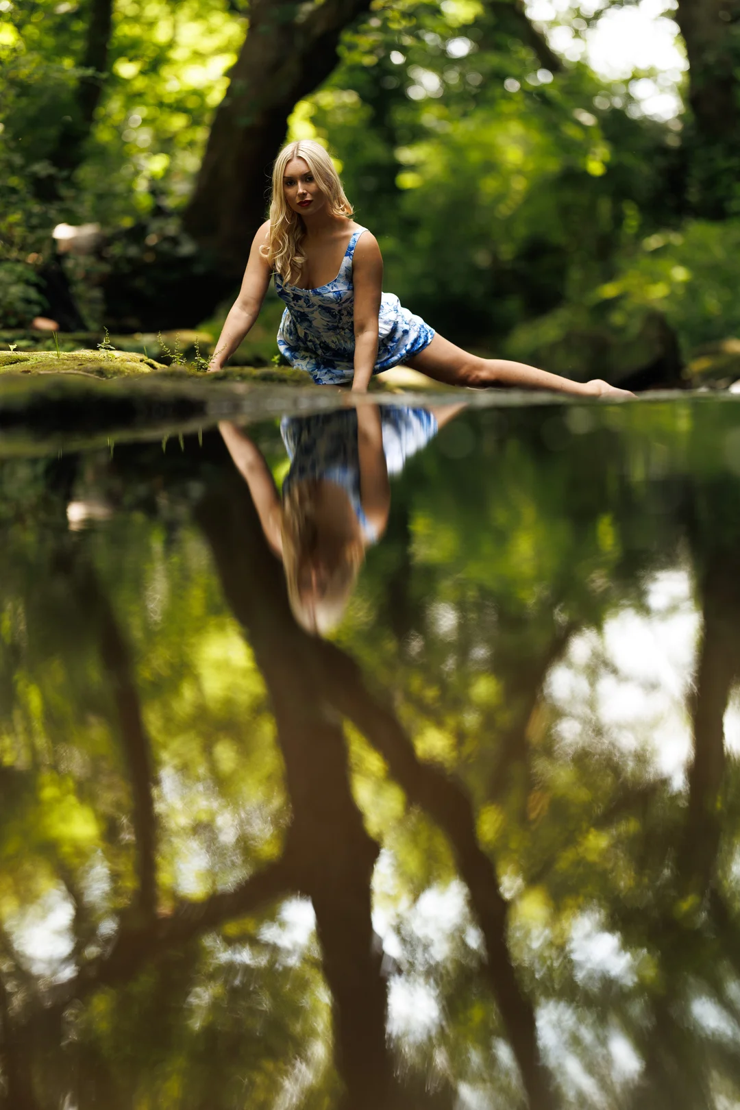 Blonde woman in a blue dress posing by a forest stream with a reflection in the water.
