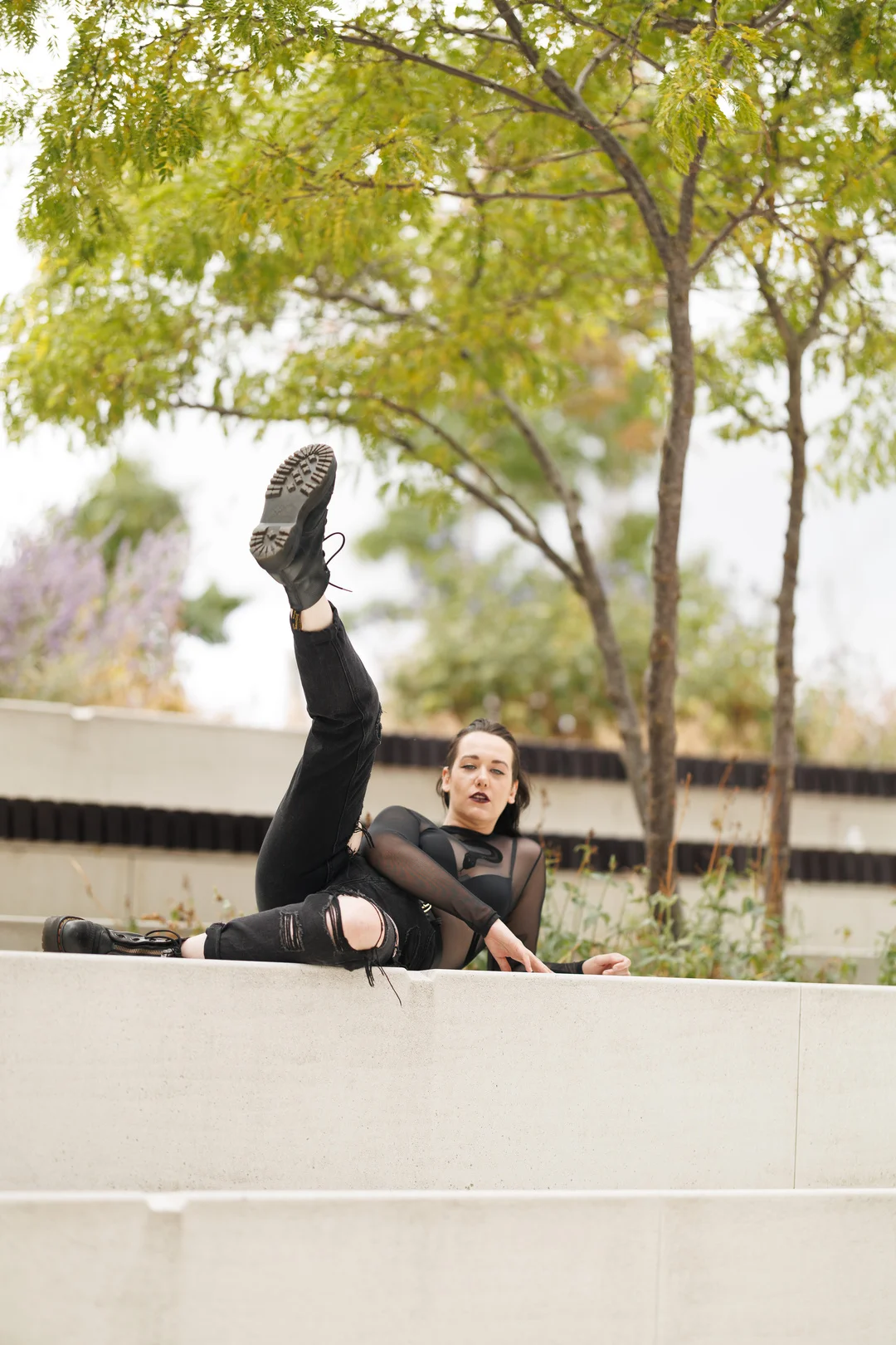 Woman in a black mesh top and ripped jeans posing on a concrete ledge with one leg raised, outdoors.