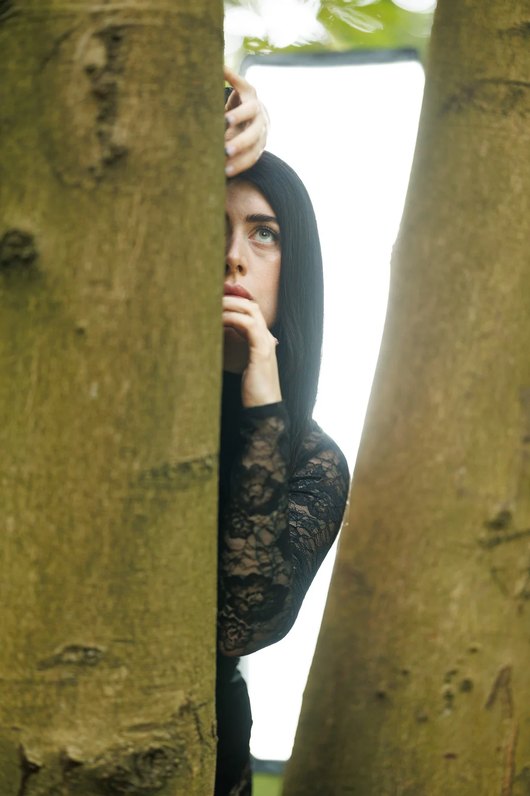 A woman with long black hair and a black lace top peeking from behind a tree trunk in a forest.