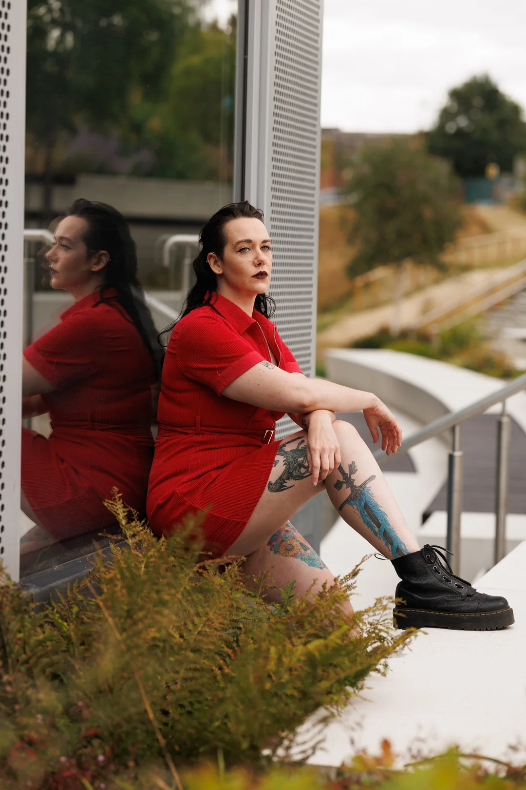 Woman in a red dress with leg tattoos sitting outdoors beside a reflective glass wall.