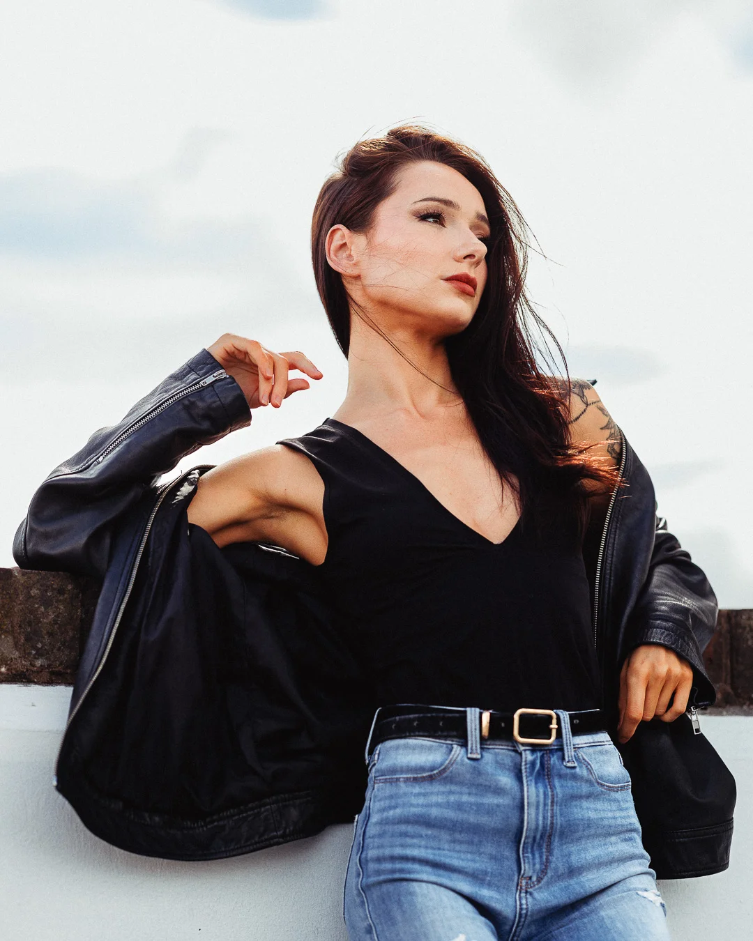 A woman with long dark hair posing outdoors in a black leather jacket, black top, and blue jeans.