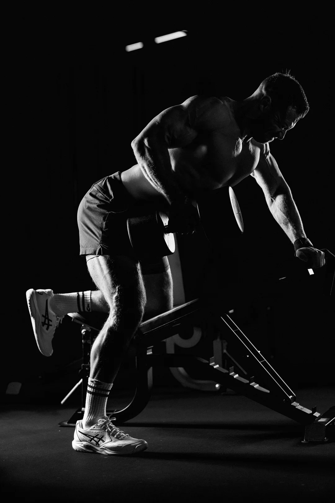 Dramatic black and white photo of a muscular man performing a dumbbell row in a gym.
