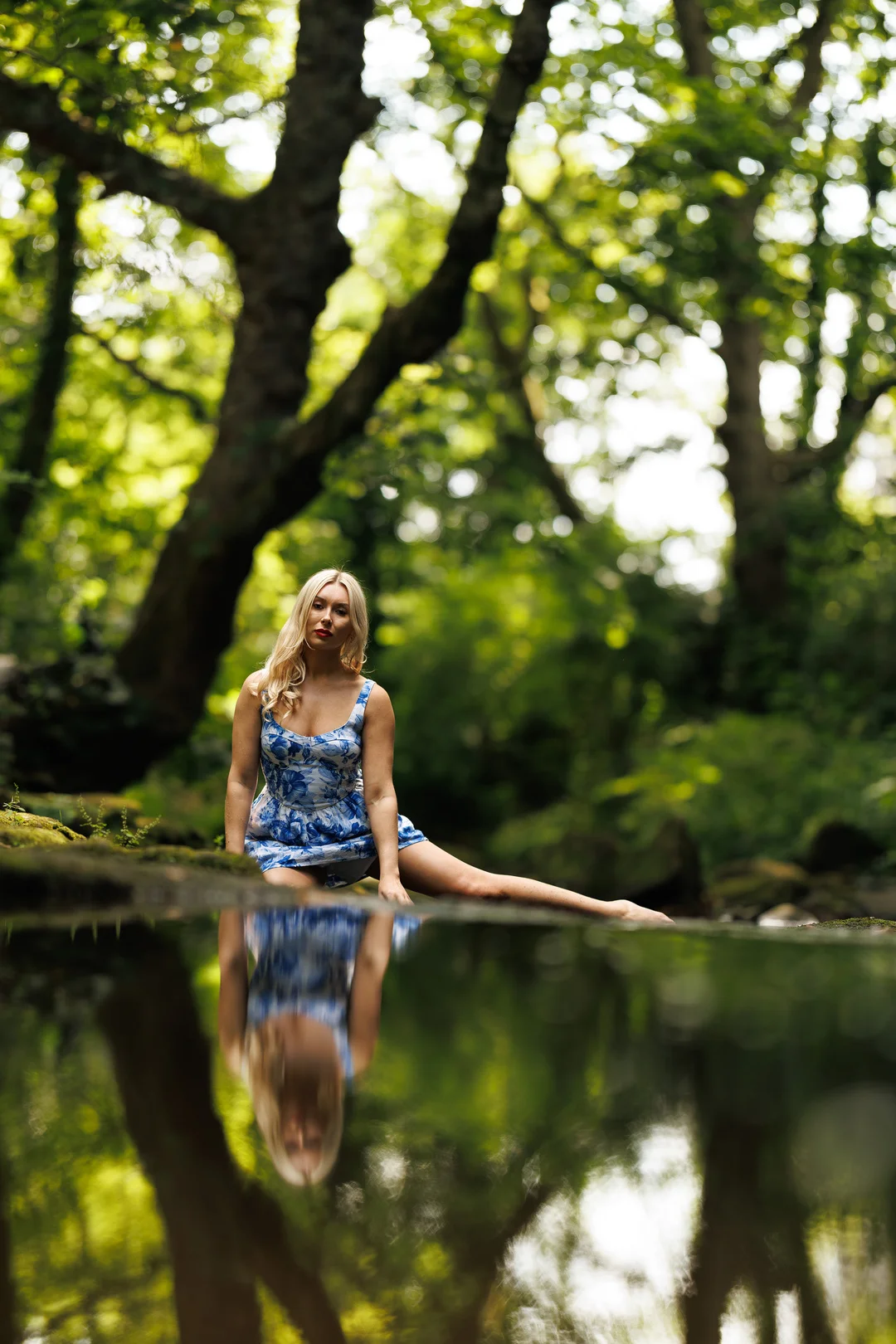 Blonde woman in a blue floral dress sitting by a reflective pond in a lush green forest.