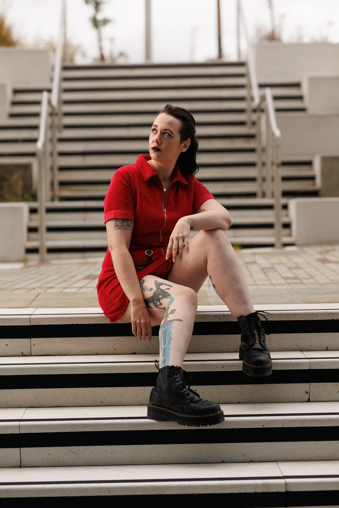 Woman with tattoos in a red zip-up dress and black boots sitting on outdoor concrete stairs.