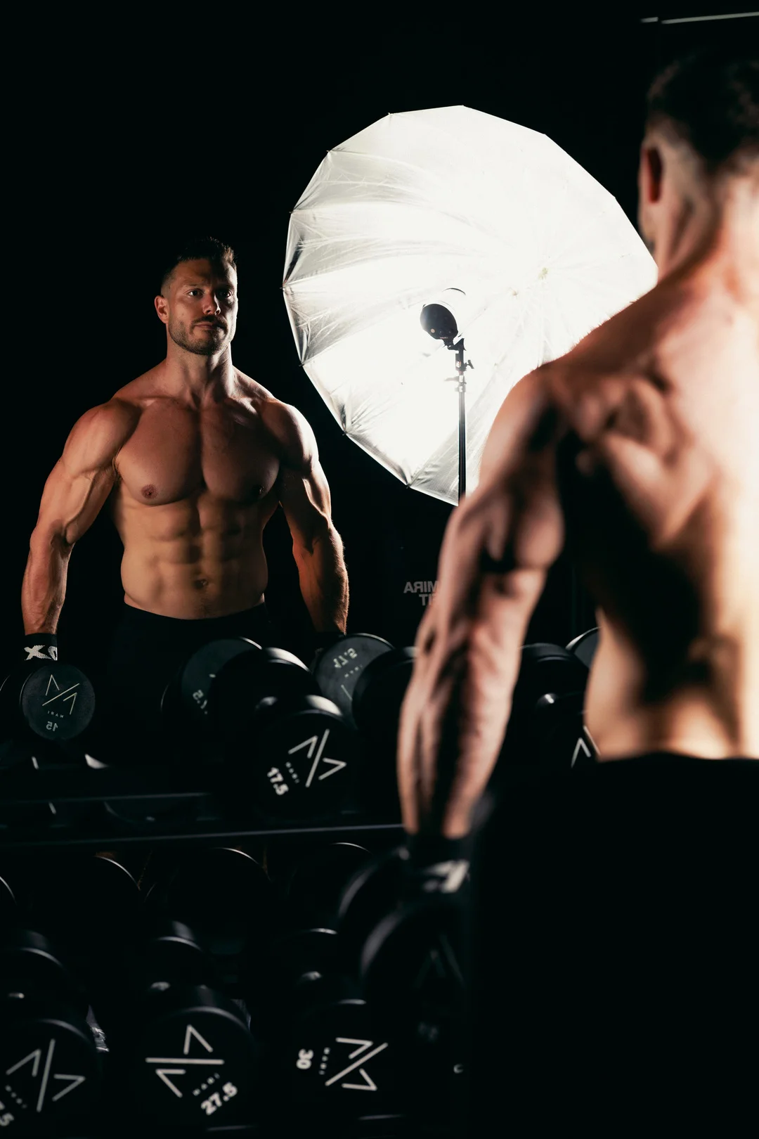Muscular man posing in a gym with studio lighting, reflected in a mirror.