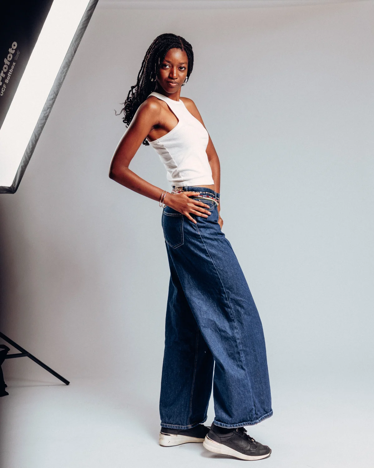 Full-body studio portrait of a Black woman with long braids wearing a white tank top and wide-leg blue jeans.