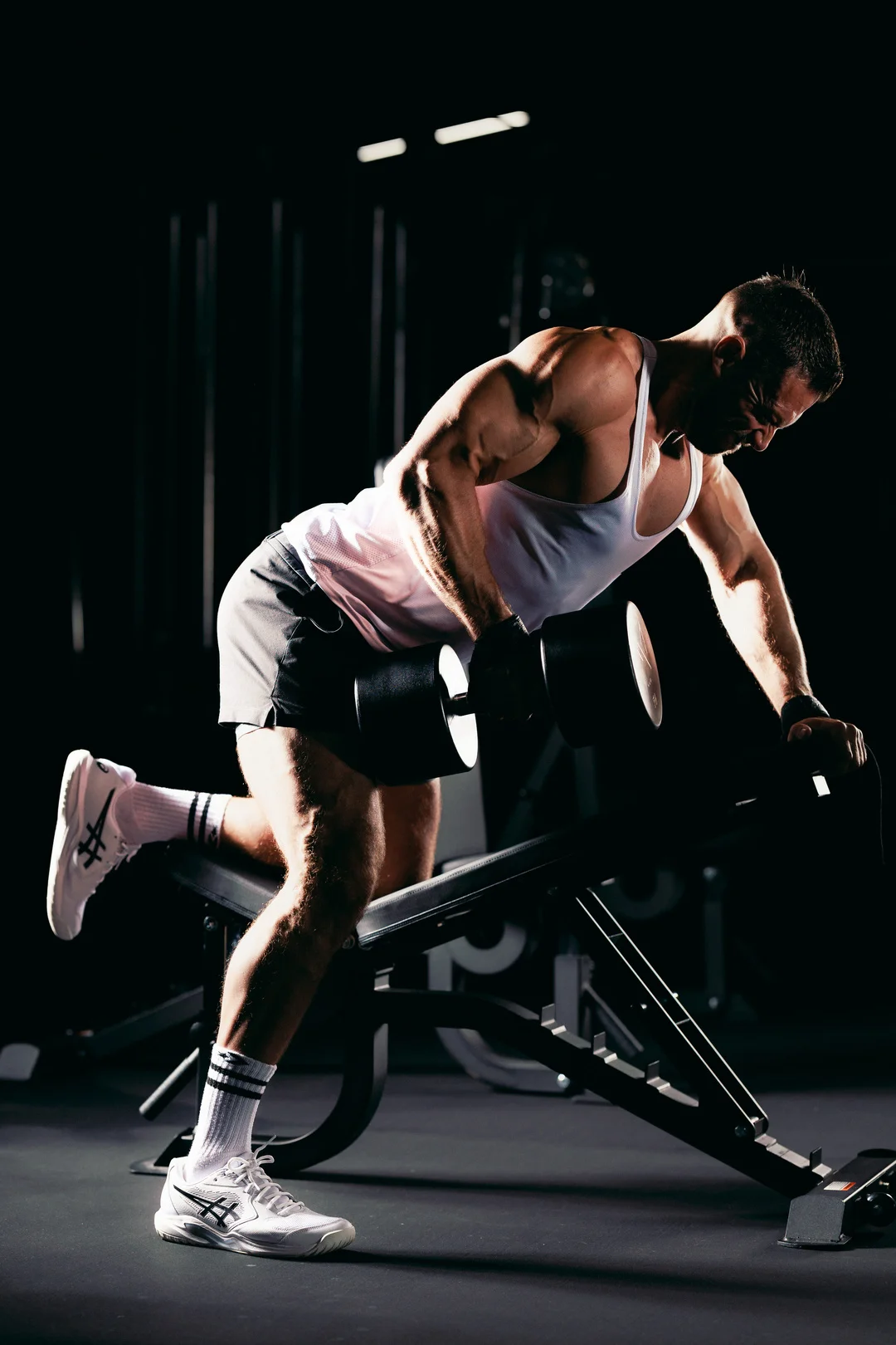 Muscular man in a white tank top performing a dumbbell row in a dark gym setting.