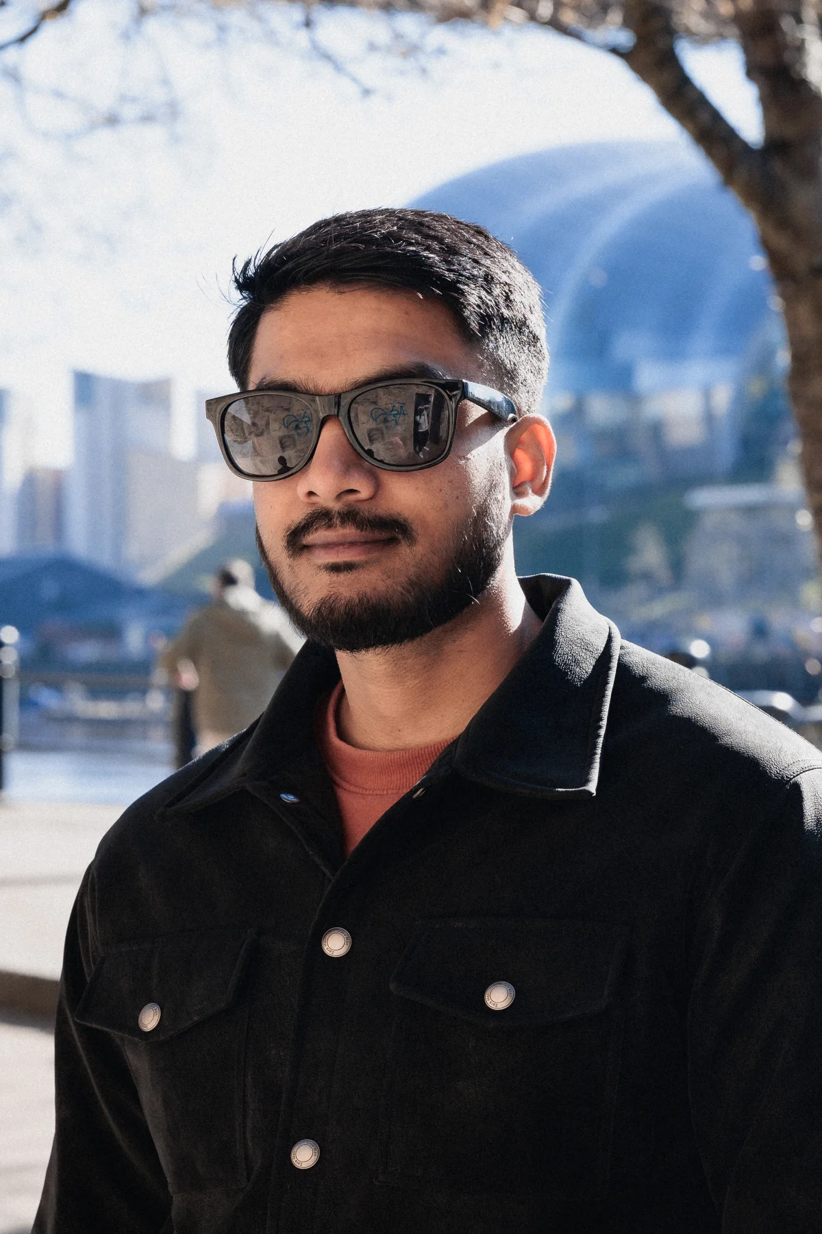 Portrait of a man with a beard and sunglasses wearing a black jacket against a blurred urban backdrop.