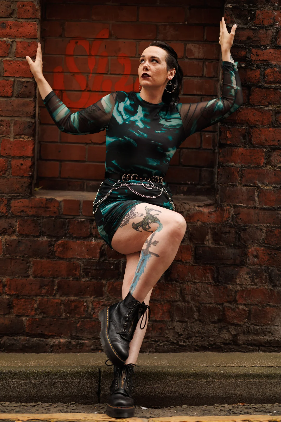 Person with tattoos and dark hair posing in a green patterned dress and black boots against a red brick wall.