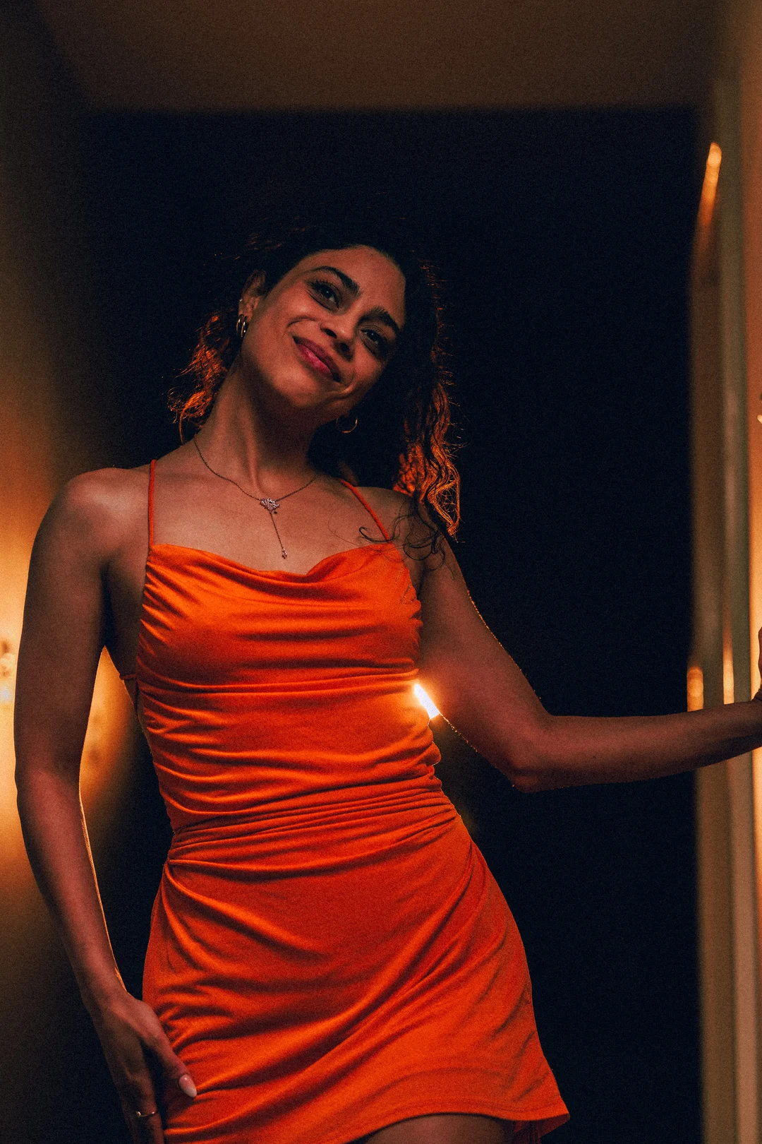 A woman with curly hair in a bright orange satin dress posing in a dimly lit room.