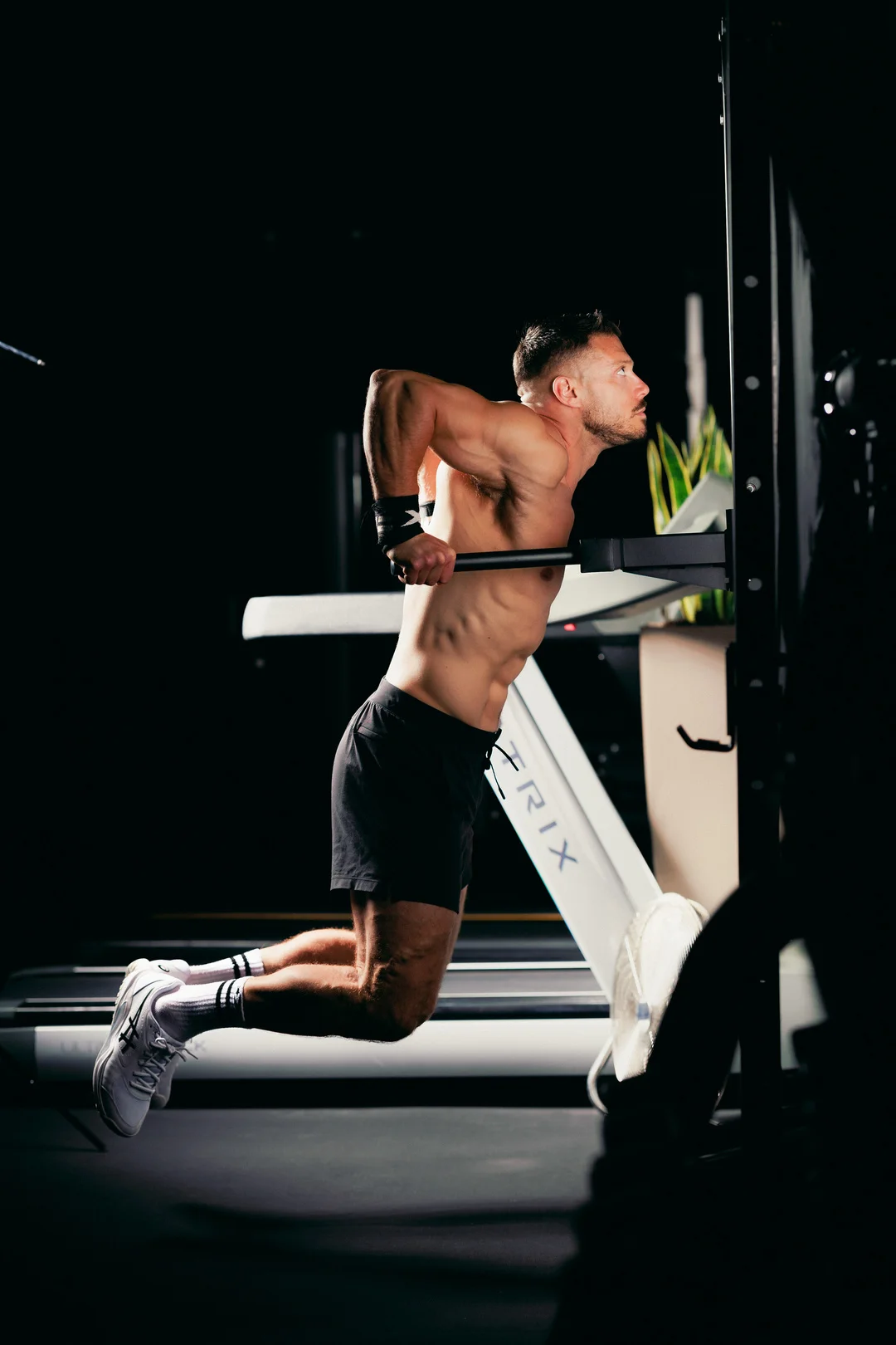 Shirtless man in black shorts performing a dip exercise on gym equipment with dramatic lighting.