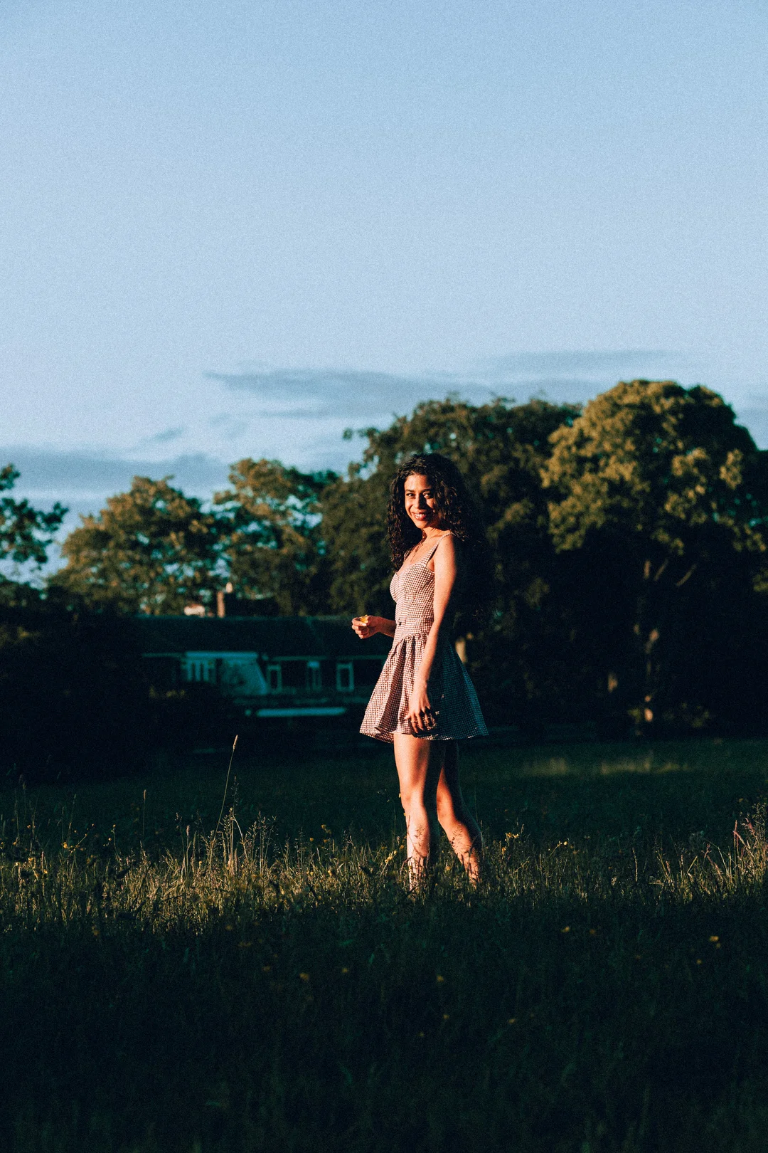 A young woman with curly hair in a red and white checkered dress standing in a grassy field during golden hour.