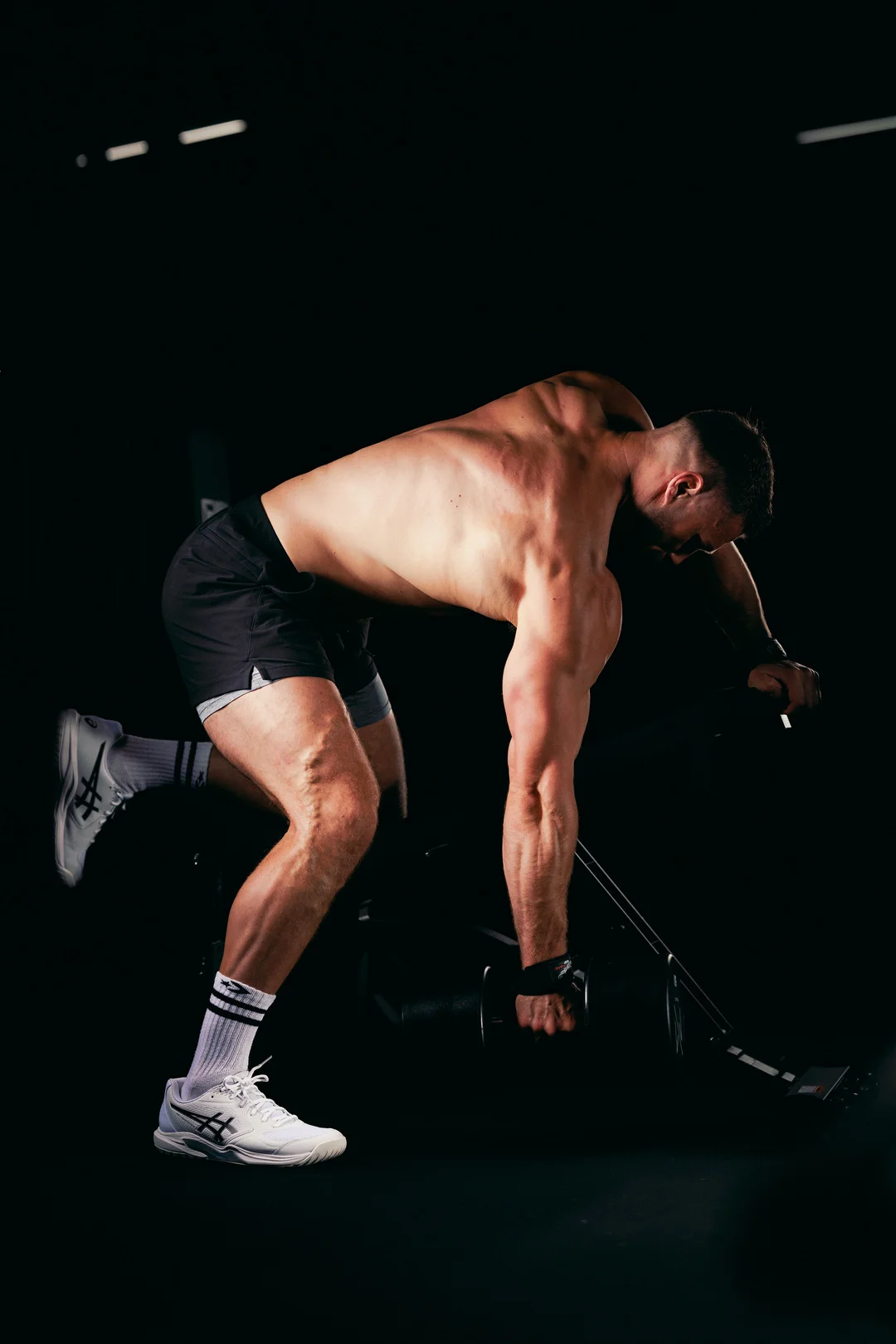 Shirtless man performing a dumbbell row in a dark gym with dramatic lighting.