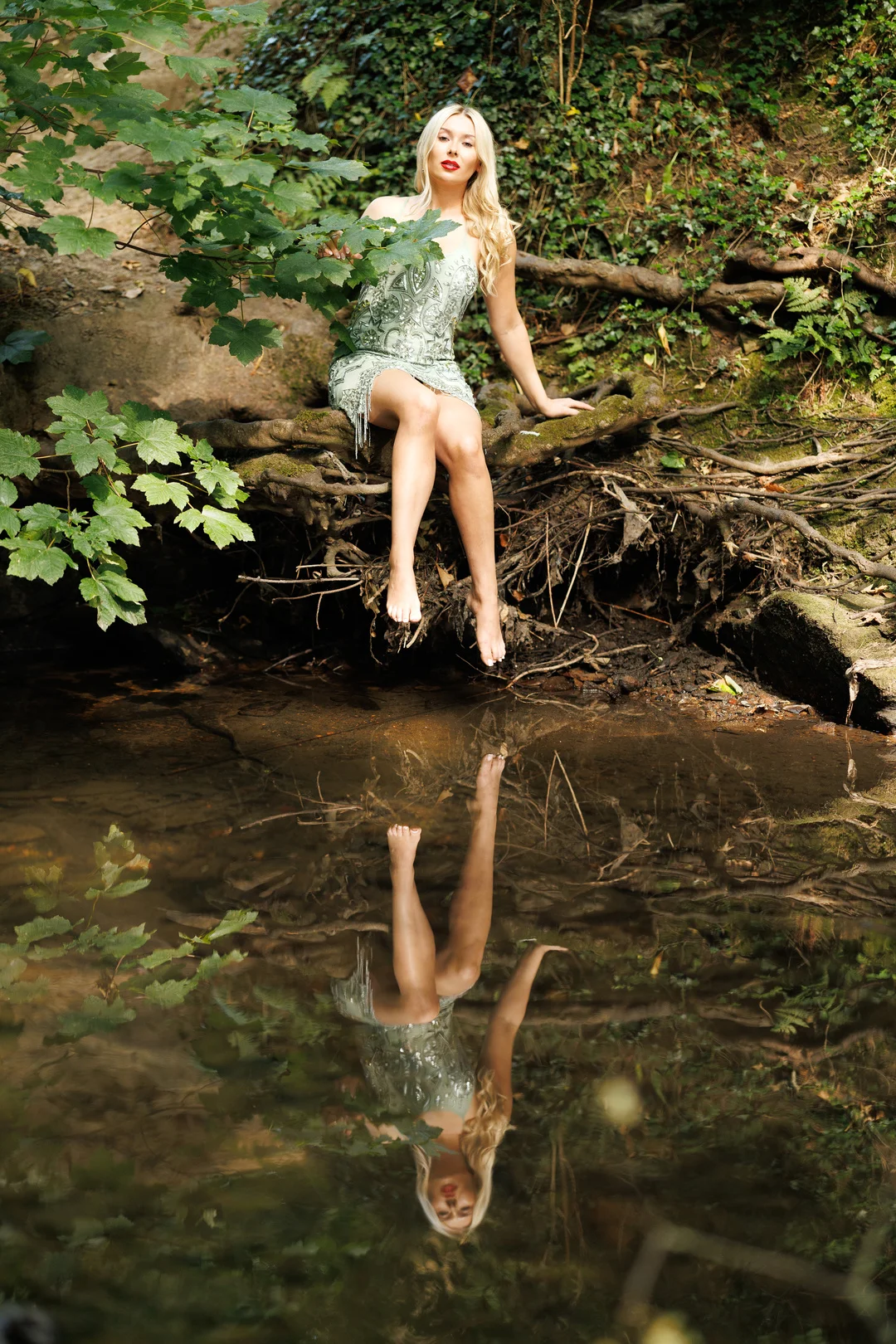 Blonde woman in a sequined green dress sitting on a riverbank with a clear reflection in the water.