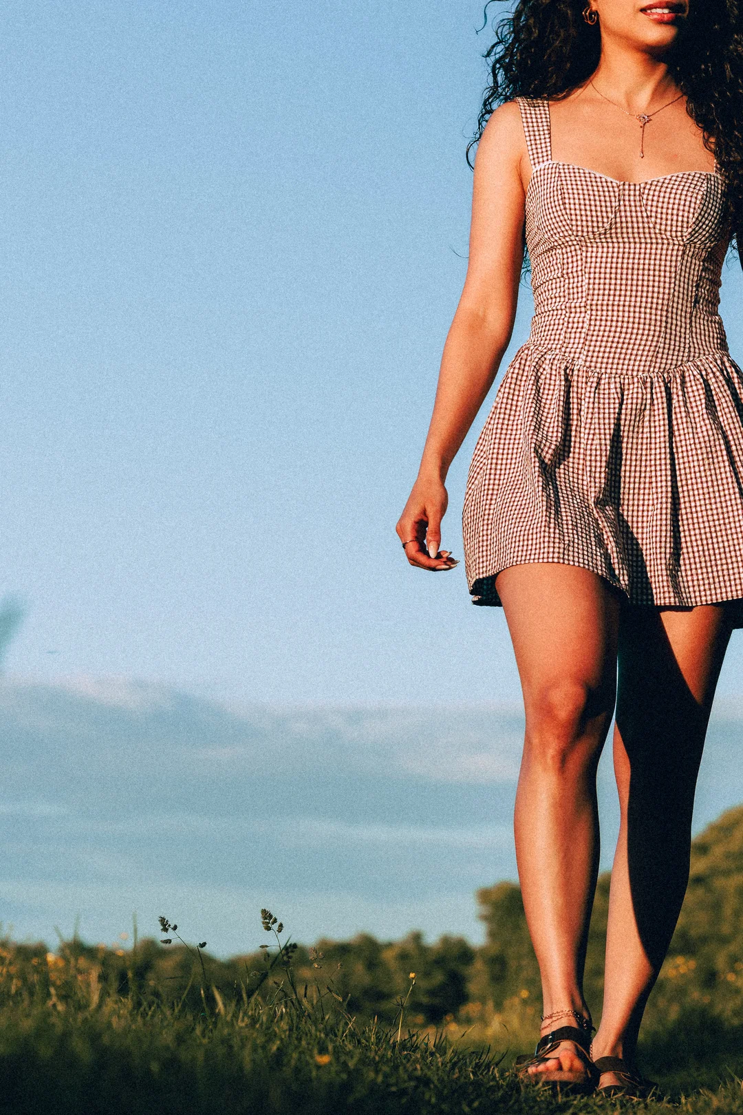 Woman in a red and white checkered mini dress standing in a grassy field under a clear blue sky.
