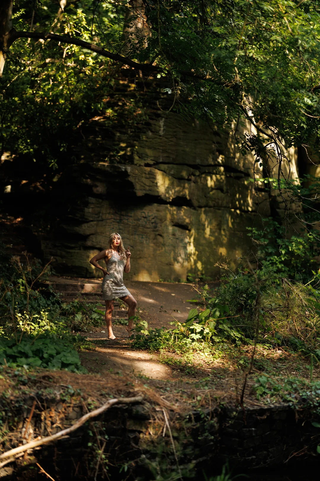 A woman in a short, sparkly dress posing against a rocky cliff in a sun-dappled forest.