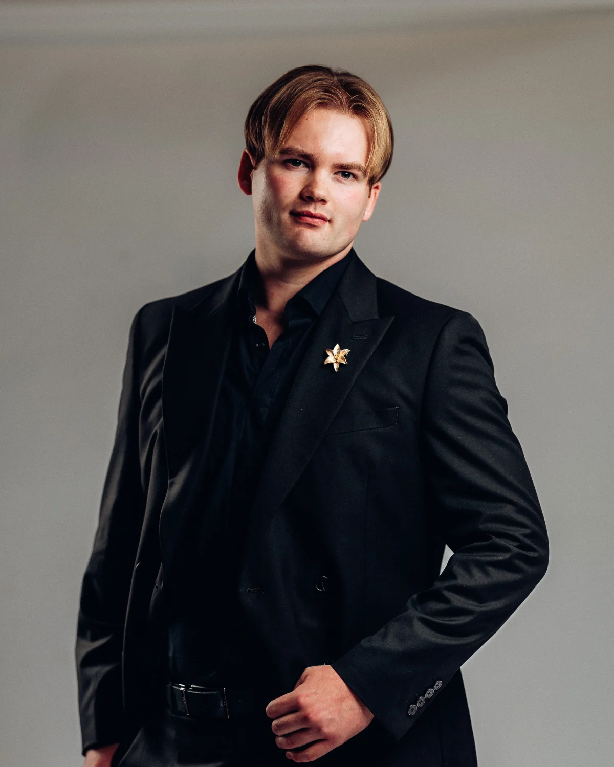 A man with reddish-blonde hair in a black suit and gold star lapel pin posing against a neutral grey background.
